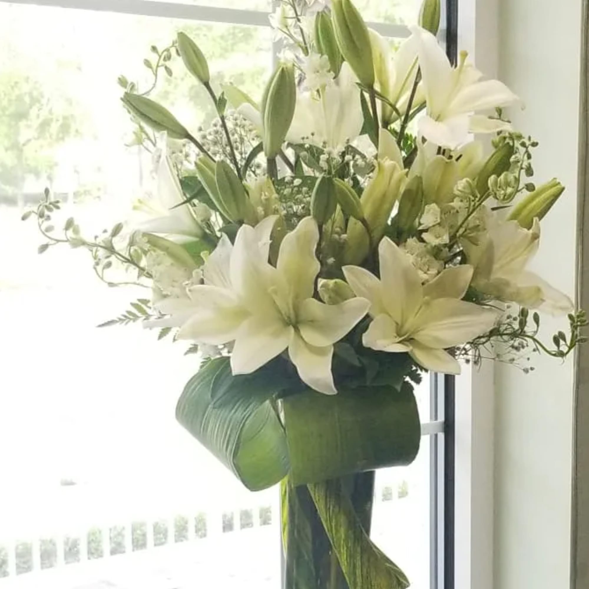 Tall arrangement of white lilies in a clear glass vase wrapped with large green leaves