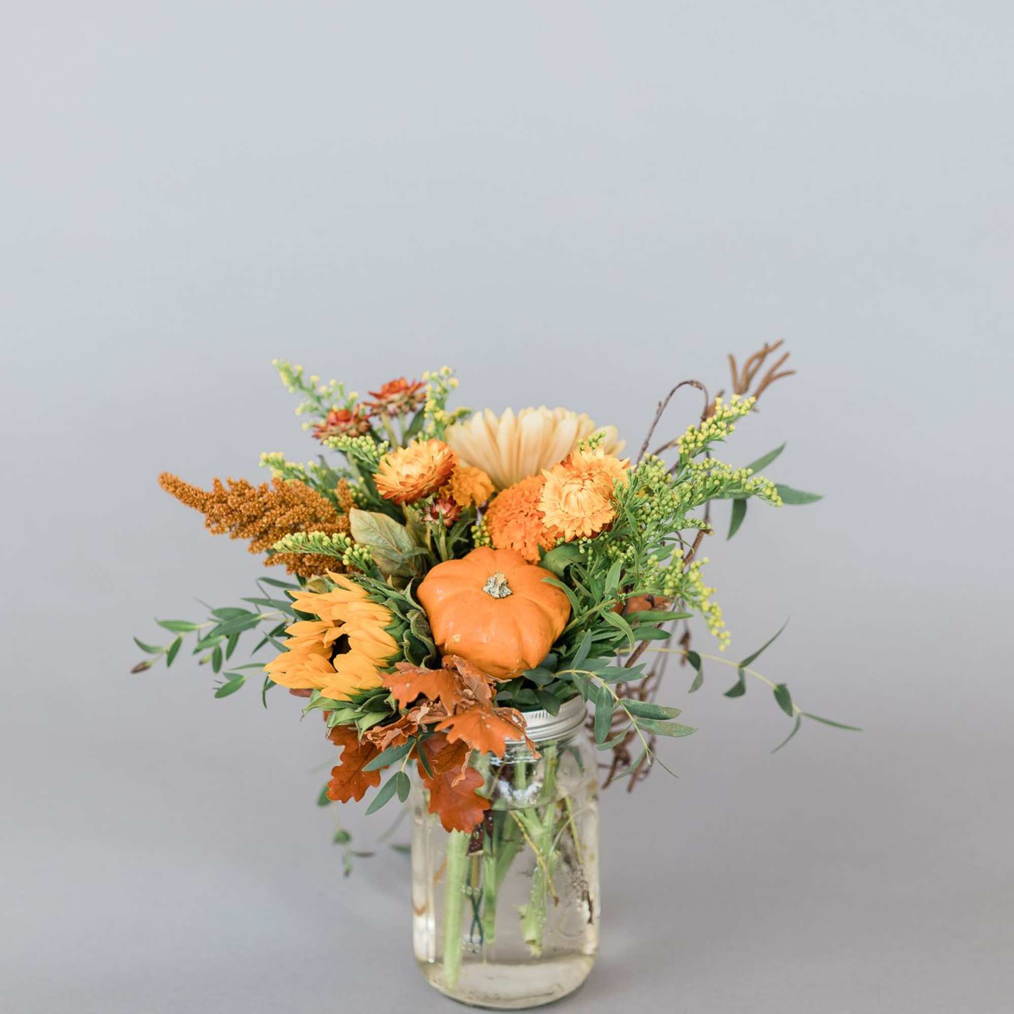 Autumn-themed bouquet of orange flowers and a small pumpkin in a clear glass mason jar