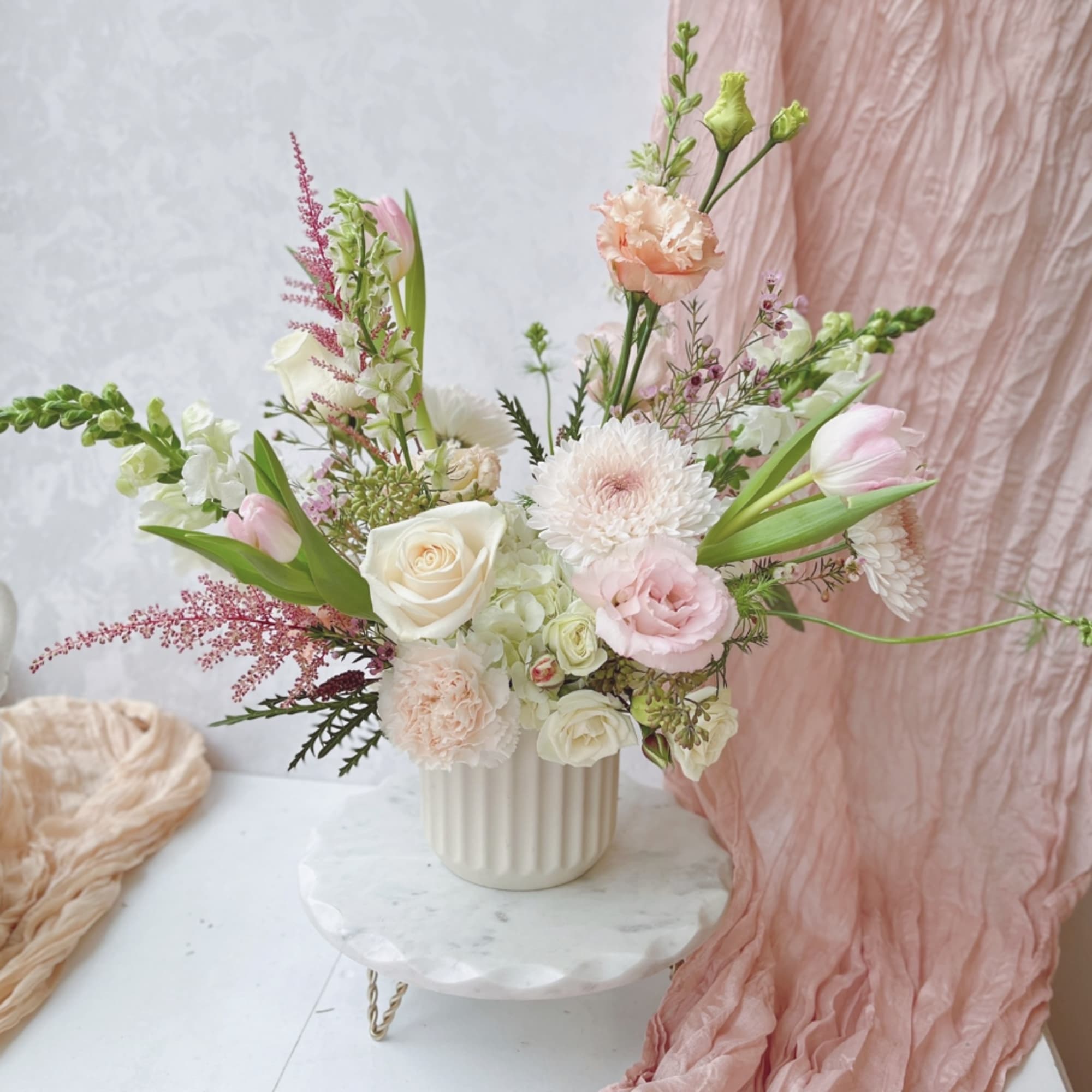 Pastel arrangement of pink and cream flowers in a white ceramic pot on a small marble stand.