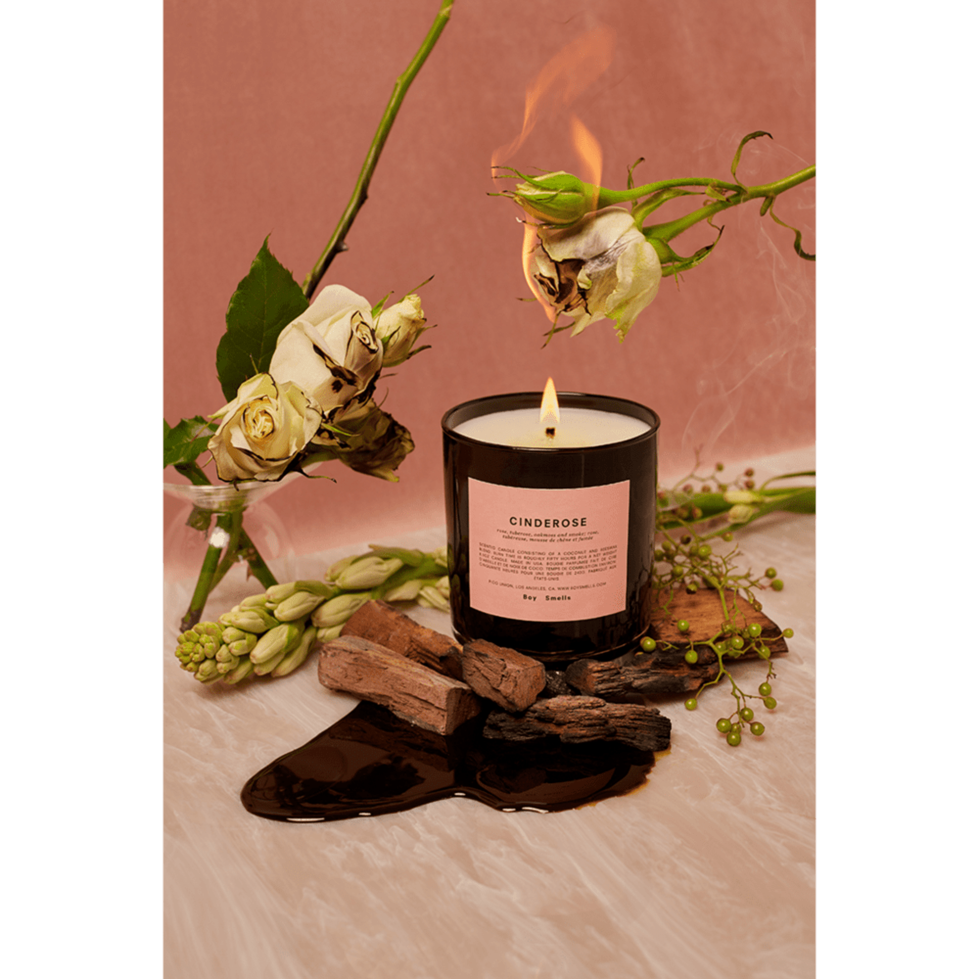 Black glass candle with label, surrounded by roses, wood pieces, and a burning rose stem