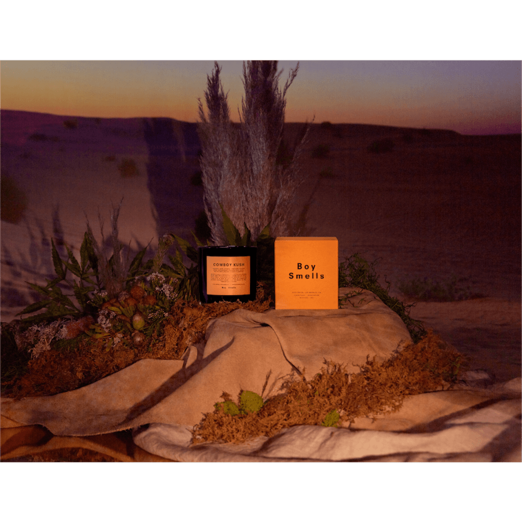 Scented candle and box displayed on moss and fabric with a desert sunset backdrop