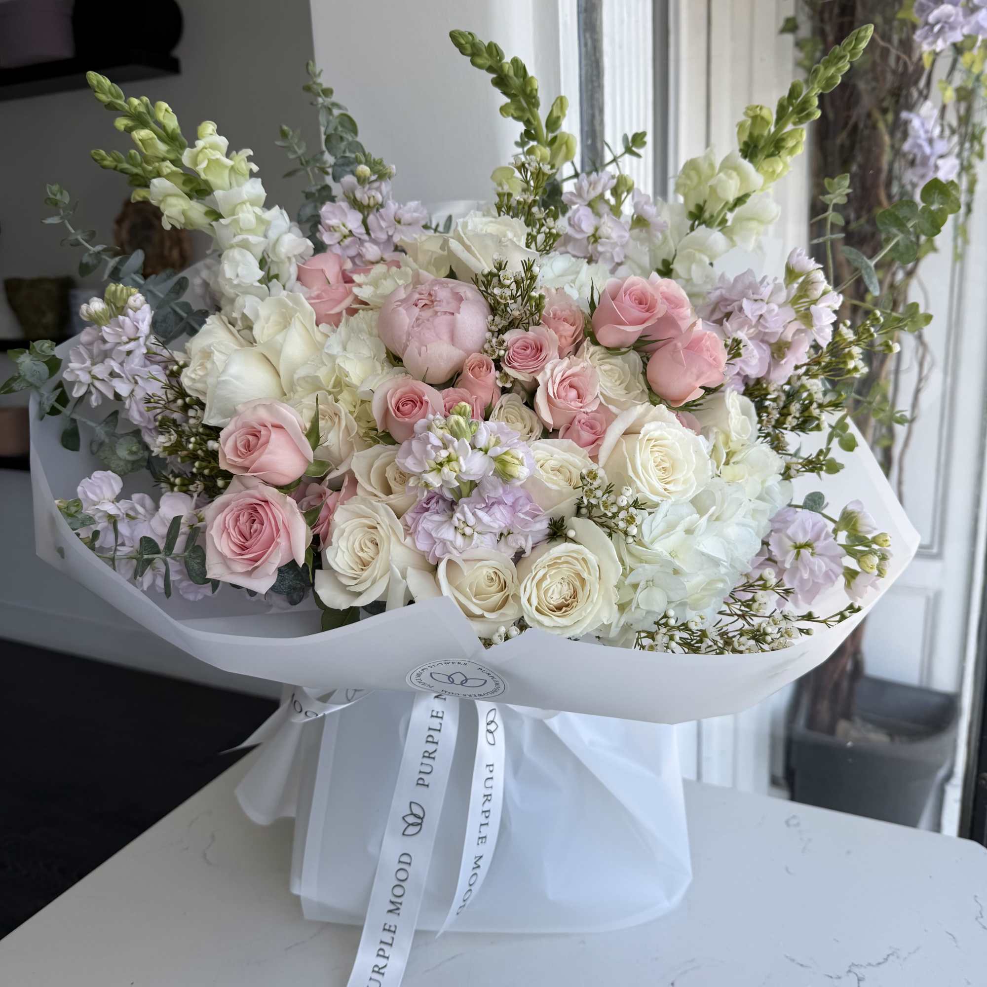 Large pastel bouquet of pink and white roses, peony, hydrangea and snapdragons wrapped in white paper with ribbon