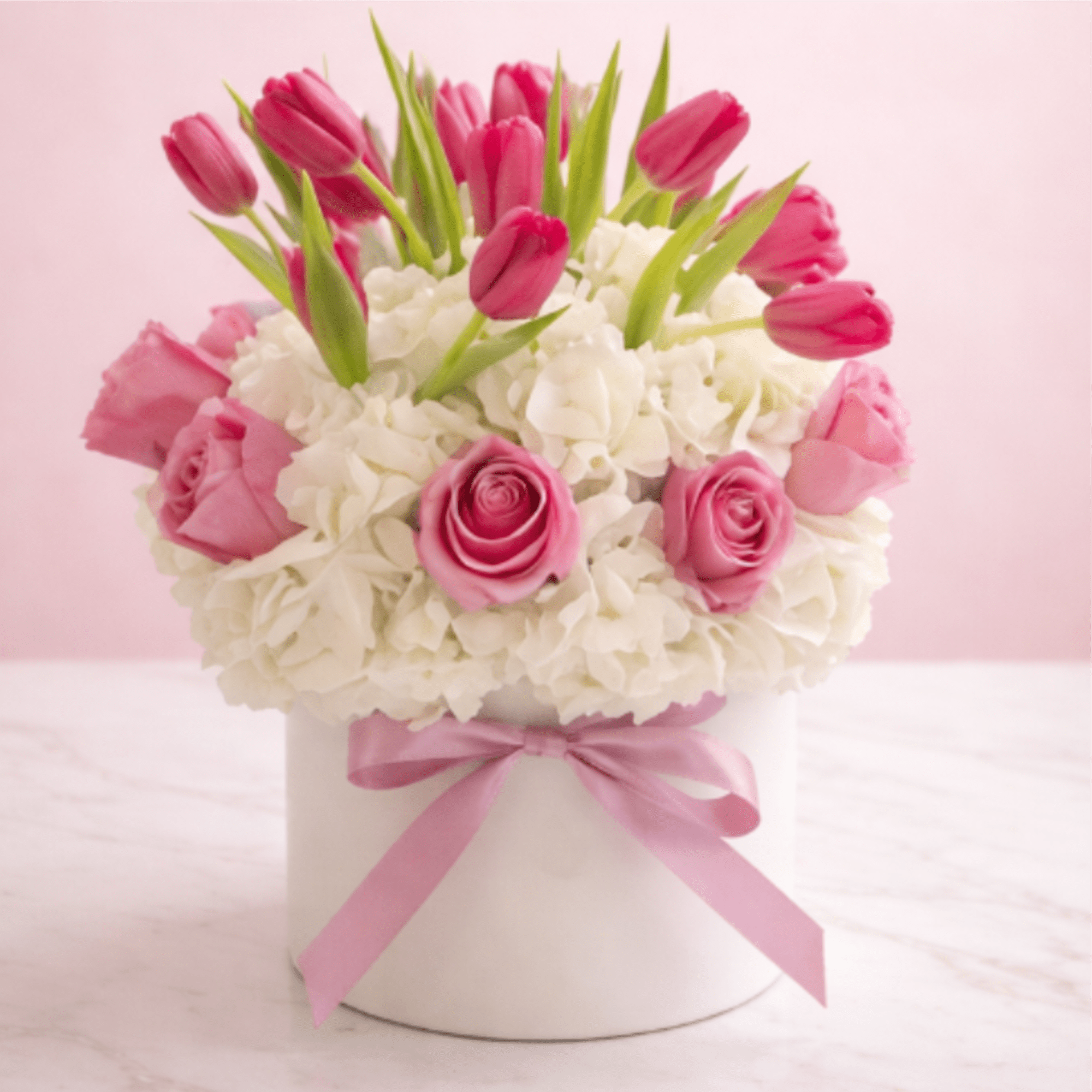 Round arrangement of pink tulips and roses set in white hydrangeas in a white hatbox with a pink ribbon.