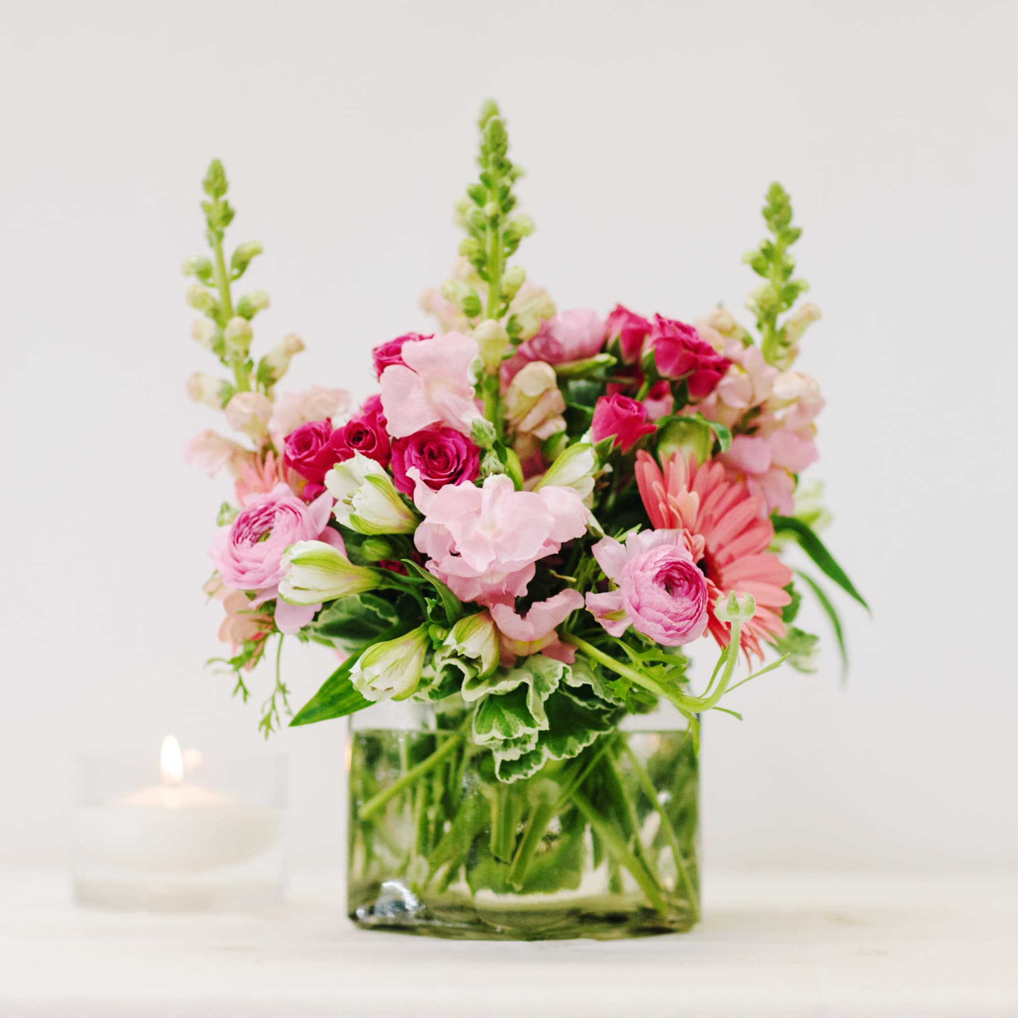 Pink mixed flowers arranged in a square glass vase beside a candle