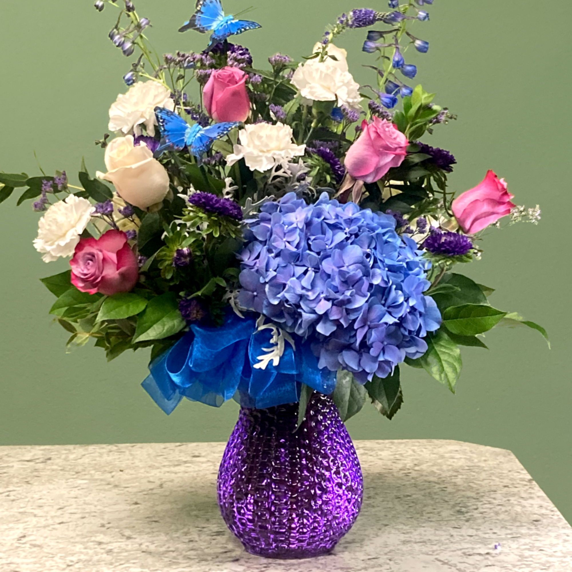 Mixed bouquet of blue hydrangea, pink roses, and white blooms in a purple glass vase
