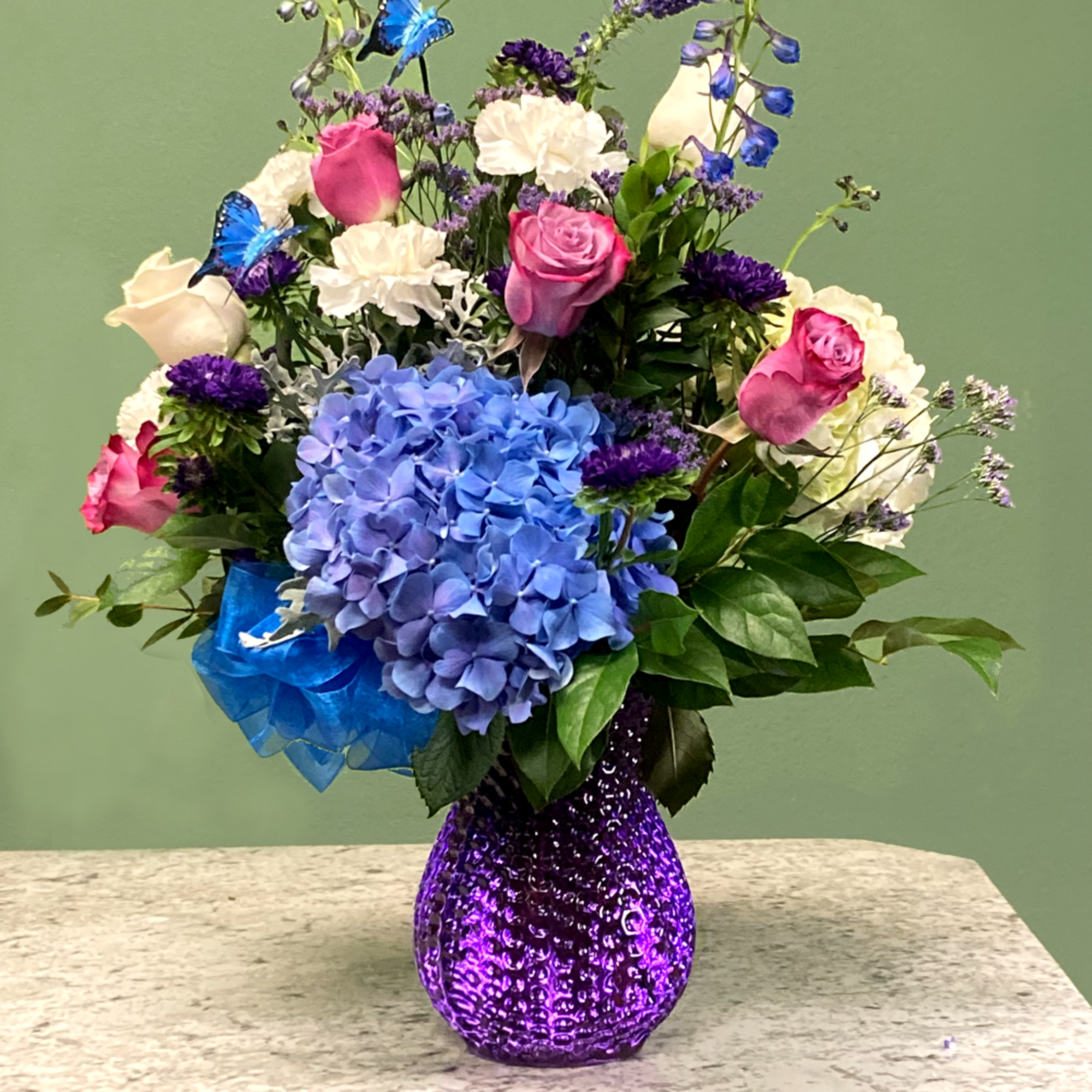 Mixed arrangement of pink roses, blue hydrangeas, and white carnations in a purple glass vase with blue butterfly accents.