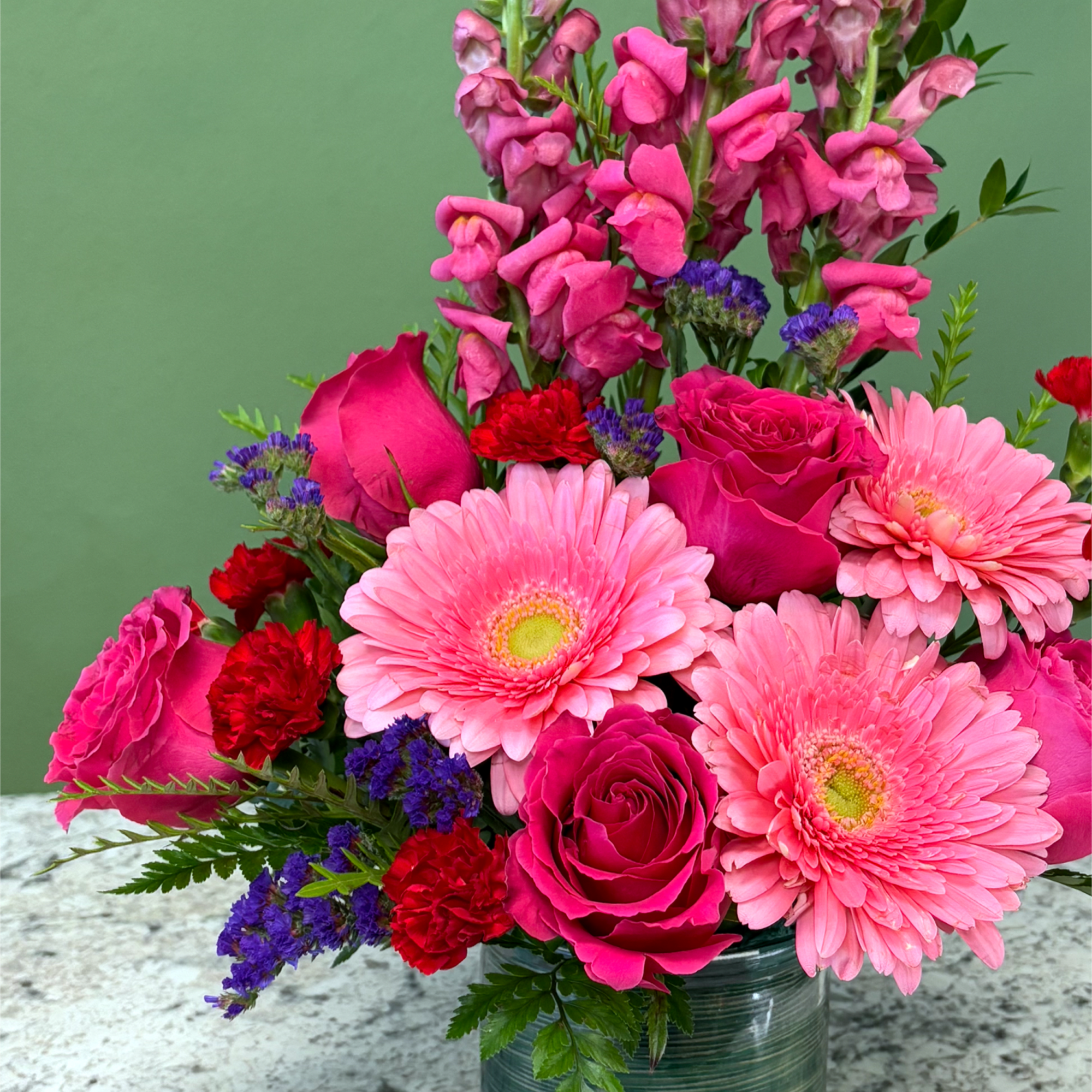 Bright arrangement of pink gerbera daisies, roses, red carnations and purple filler in a green glass vase