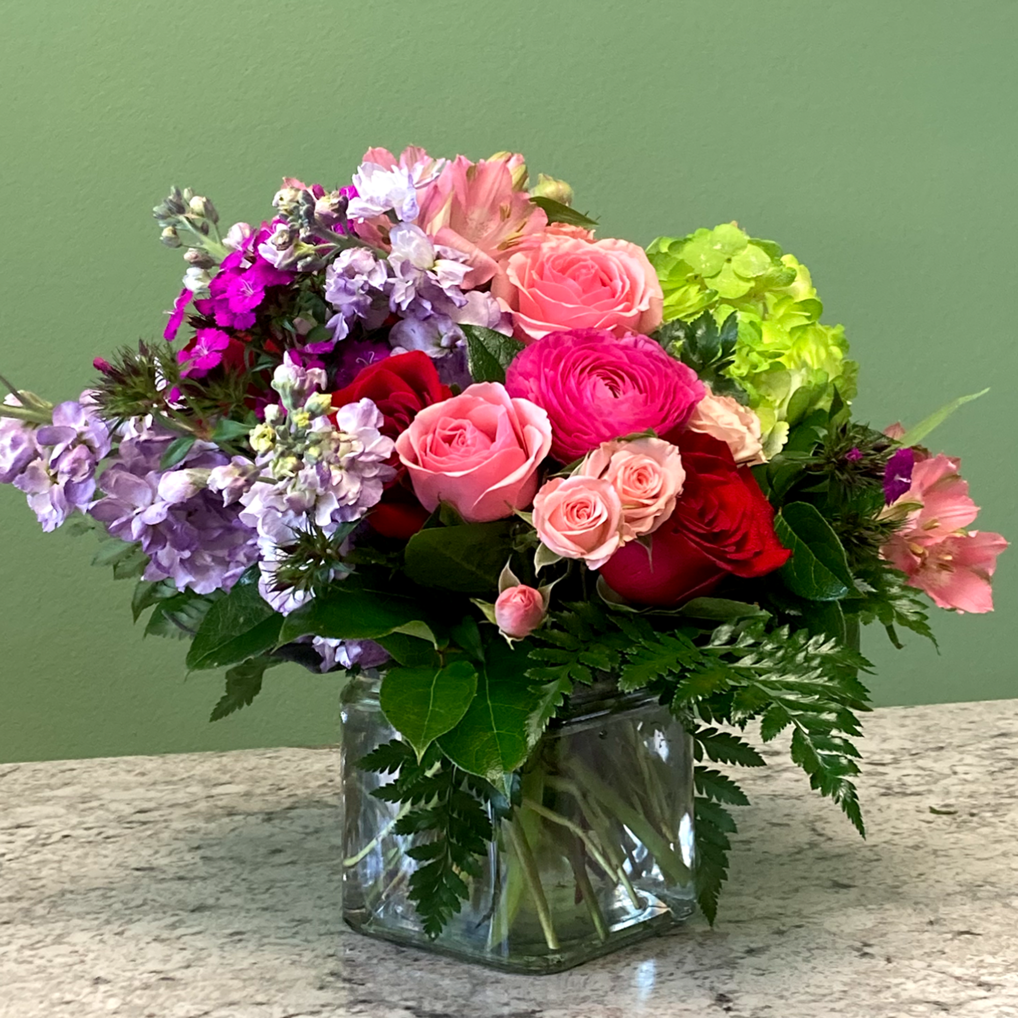 Mixed arrangement of pink and red roses with purple flowers in a clear glass cube vase