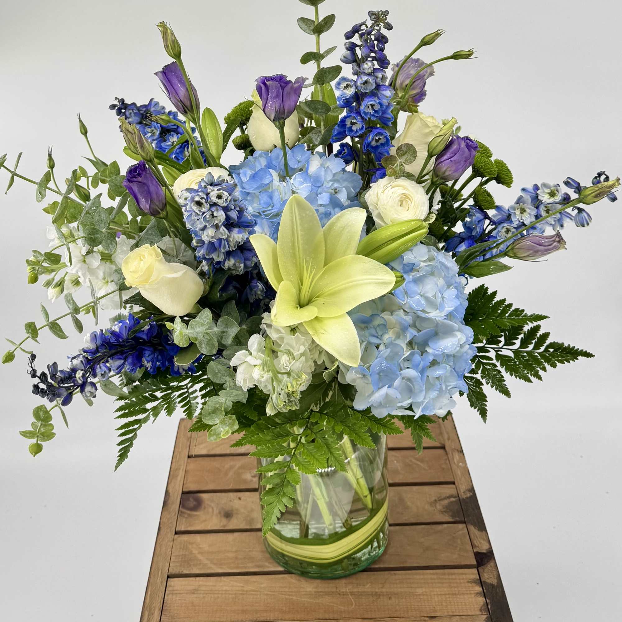 Mixed arrangement of blue hydrangeas, white roses, and yellow lilies in a clear glass vase