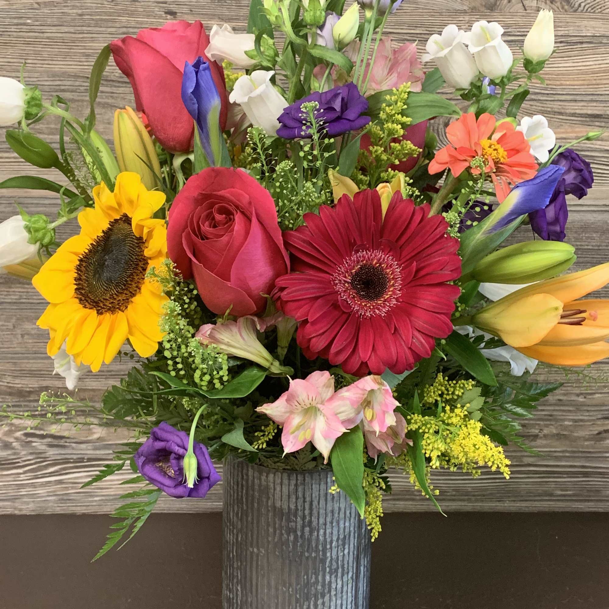 Mixed bouquet in a ribbed metal vase with roses, gerbera daisies, sunflowers, and lilies