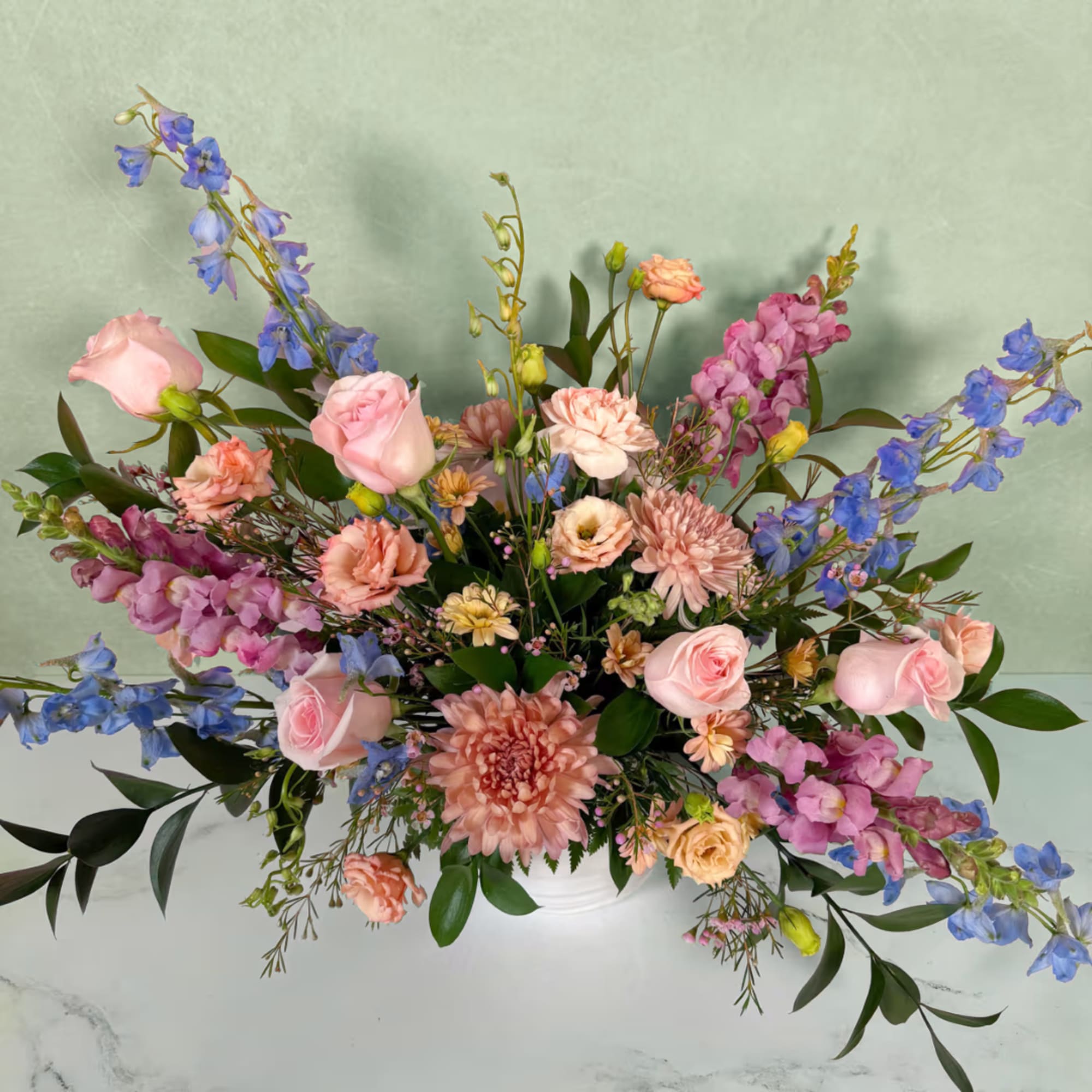 Pastel floral arrangement with pink roses, peach blooms, and blue delphinium in a white vase