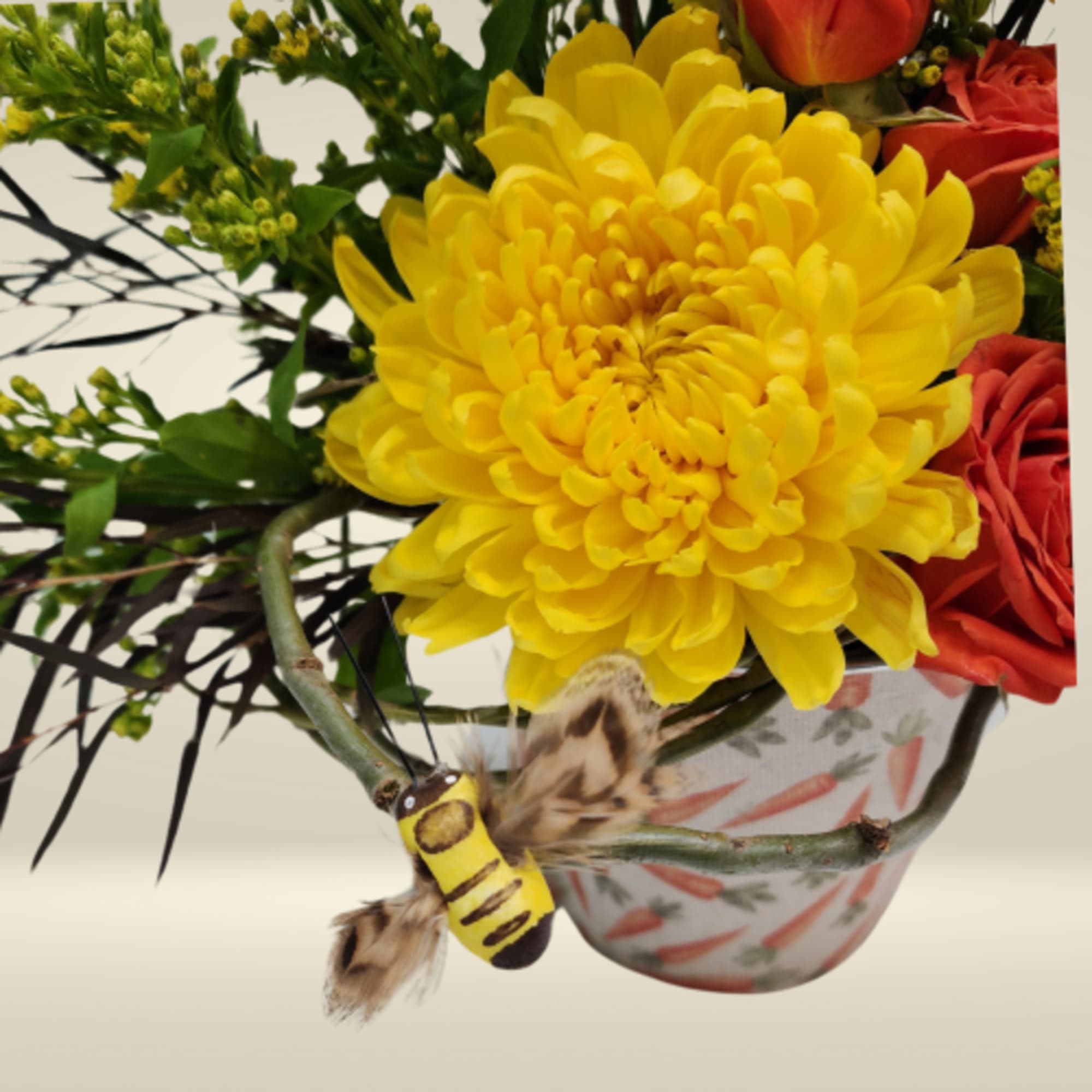 Yellow chrysanthemum and orange roses in a carrot-print container with a small decorative bee pick