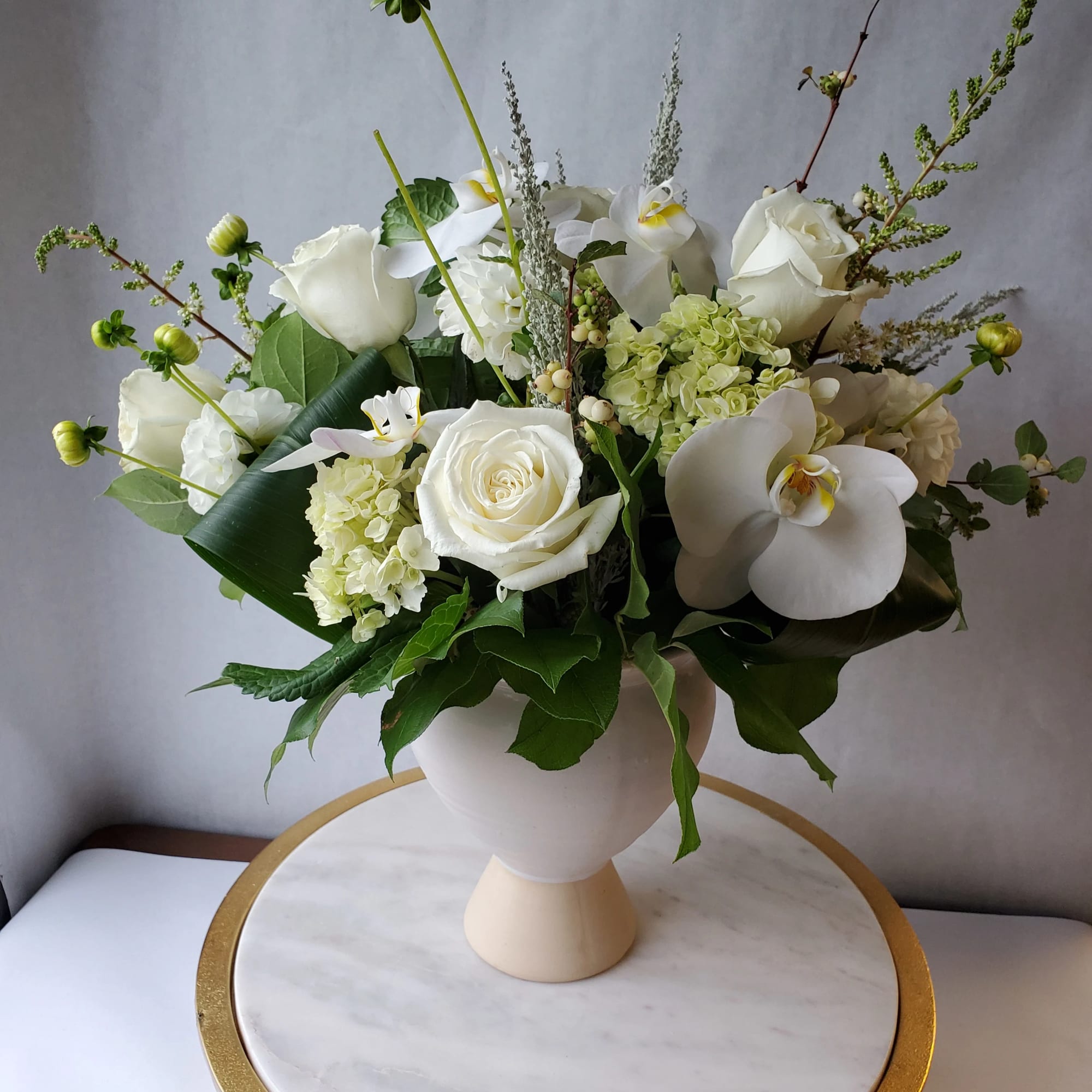 White arrangement of roses, hydrangea, and orchids in a white pedestal vase on a marble stand