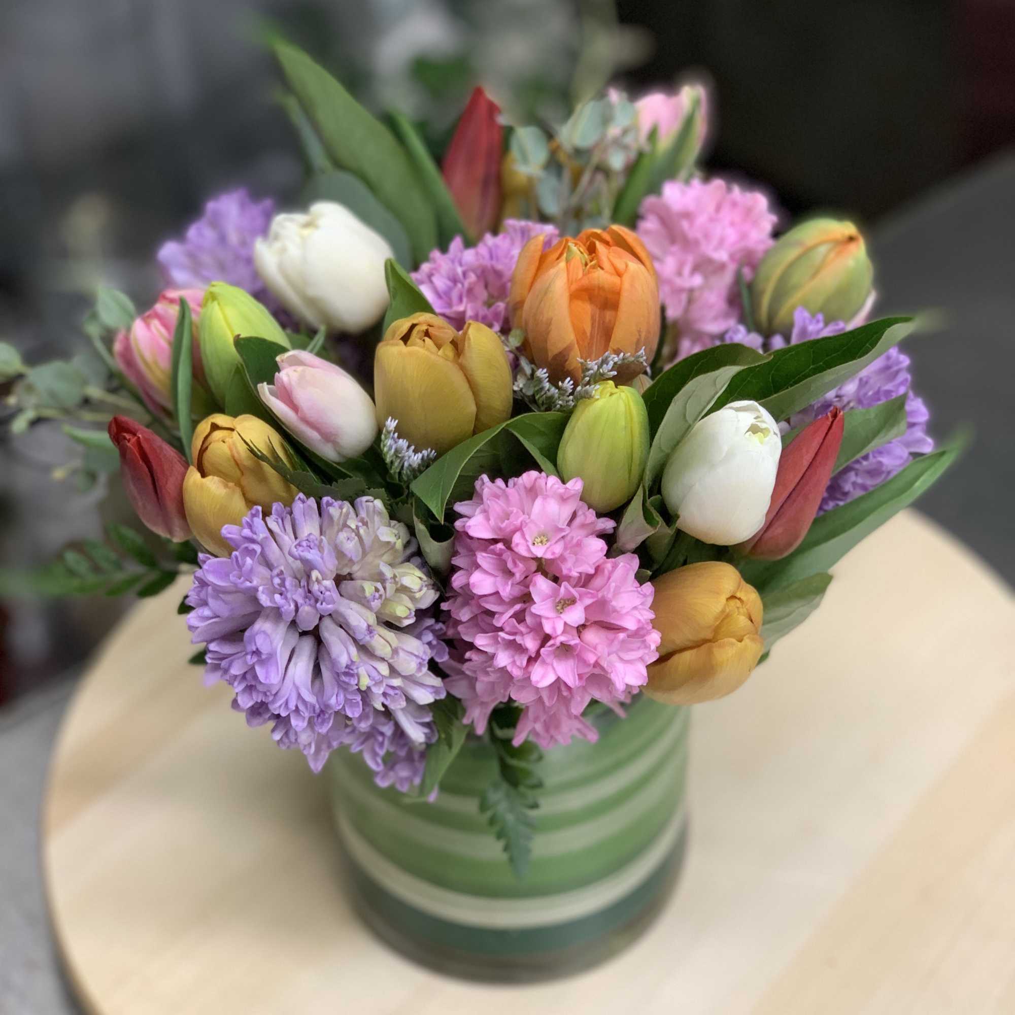Mixed tulips and pastel flowers arranged in a green striped vase on a light wood surface
