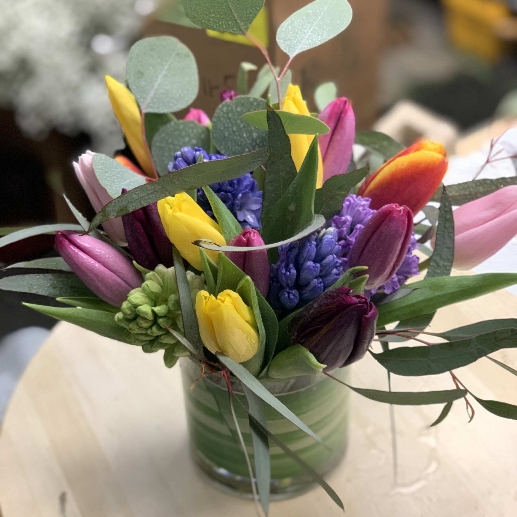 Short glass vase filled with yellow, pink, and purple tulips with purple hyacinths on a light wood table