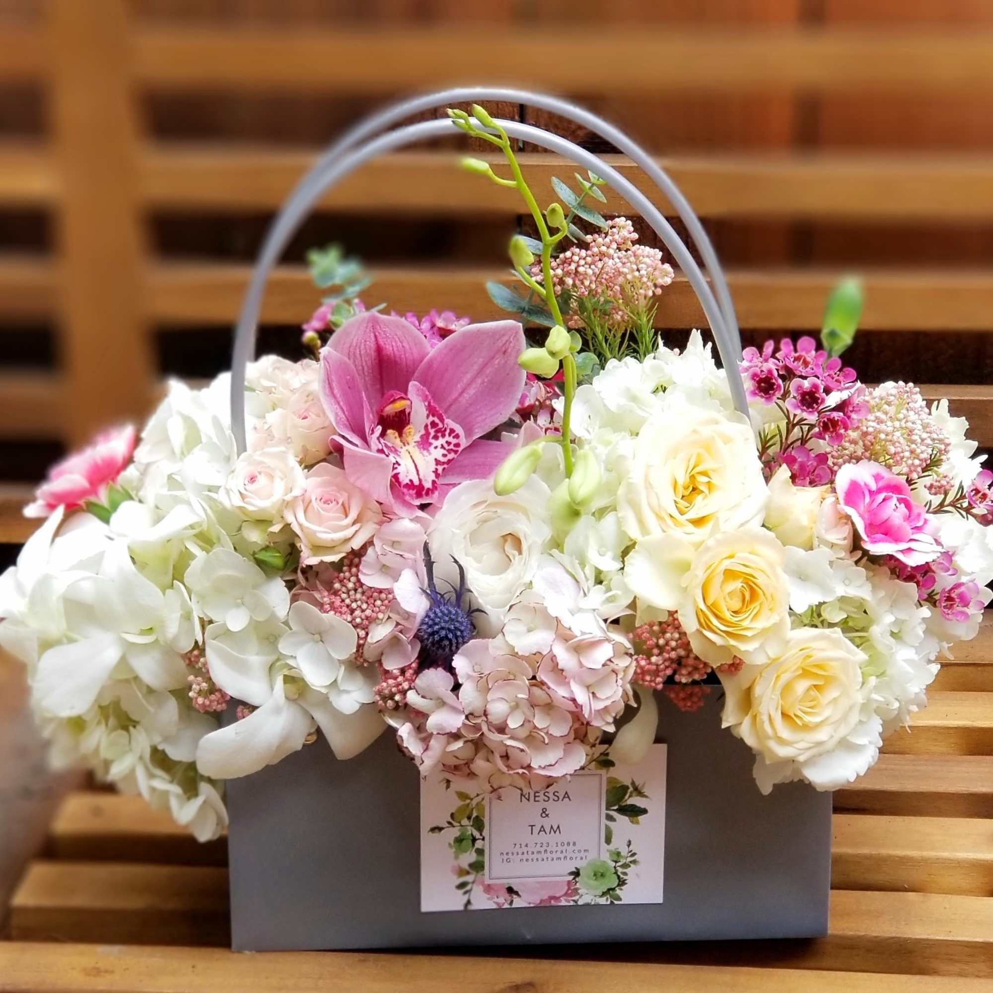 Low box arrangement of white hydrangeas, cream roses, and pink orchids in a gray handled container