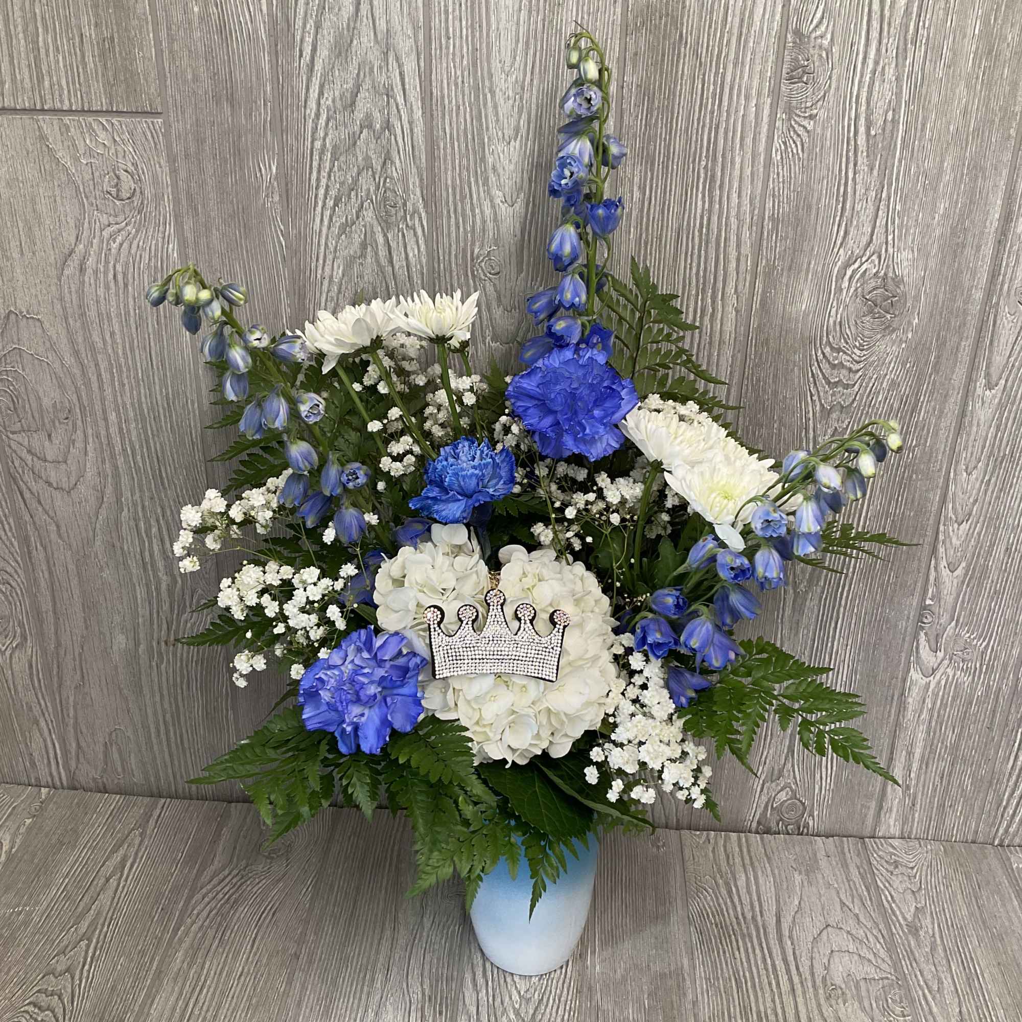Tall blue and white flower arrangement with carnations, mums, hydrangea and a crown accent in a blue vase.