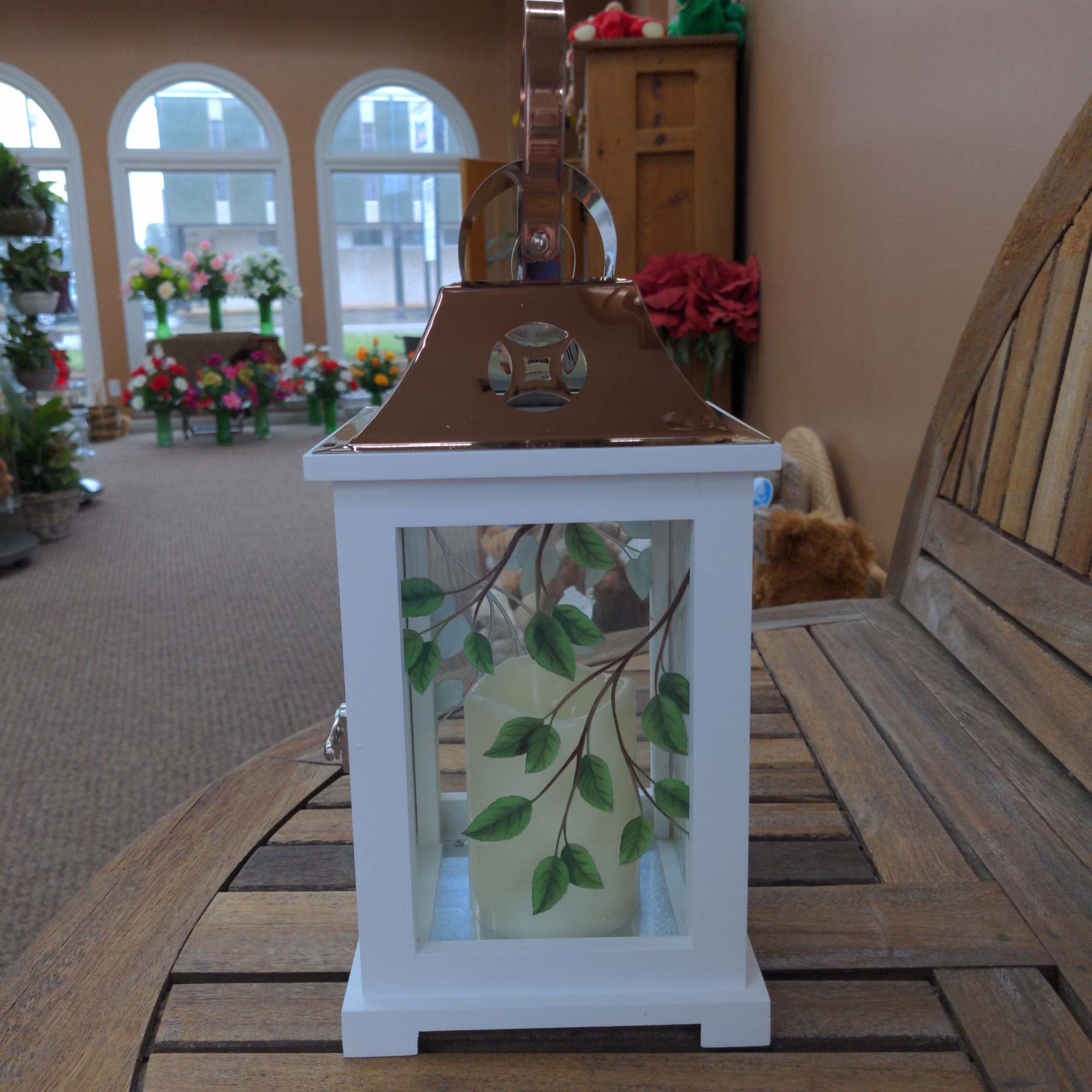 White lantern with copper top, leafy painted glass panels, and a cream pillar candle inside.