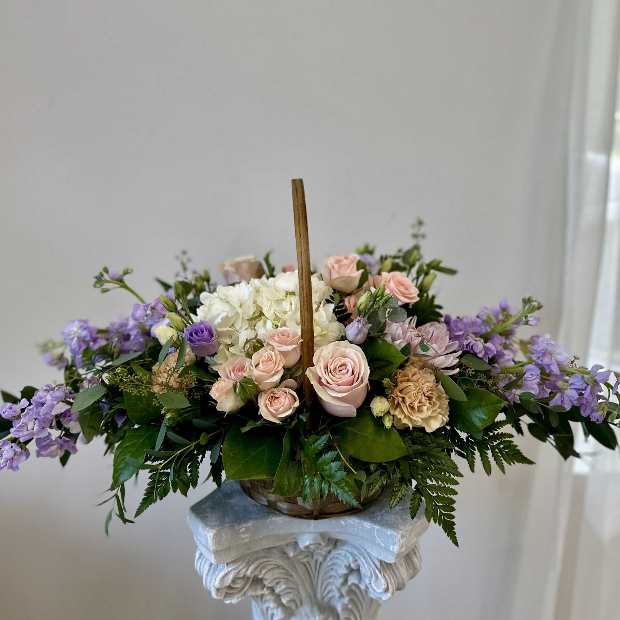 Pastel pink roses, white hydrangea, and lavender blooms arranged in a handled basket.