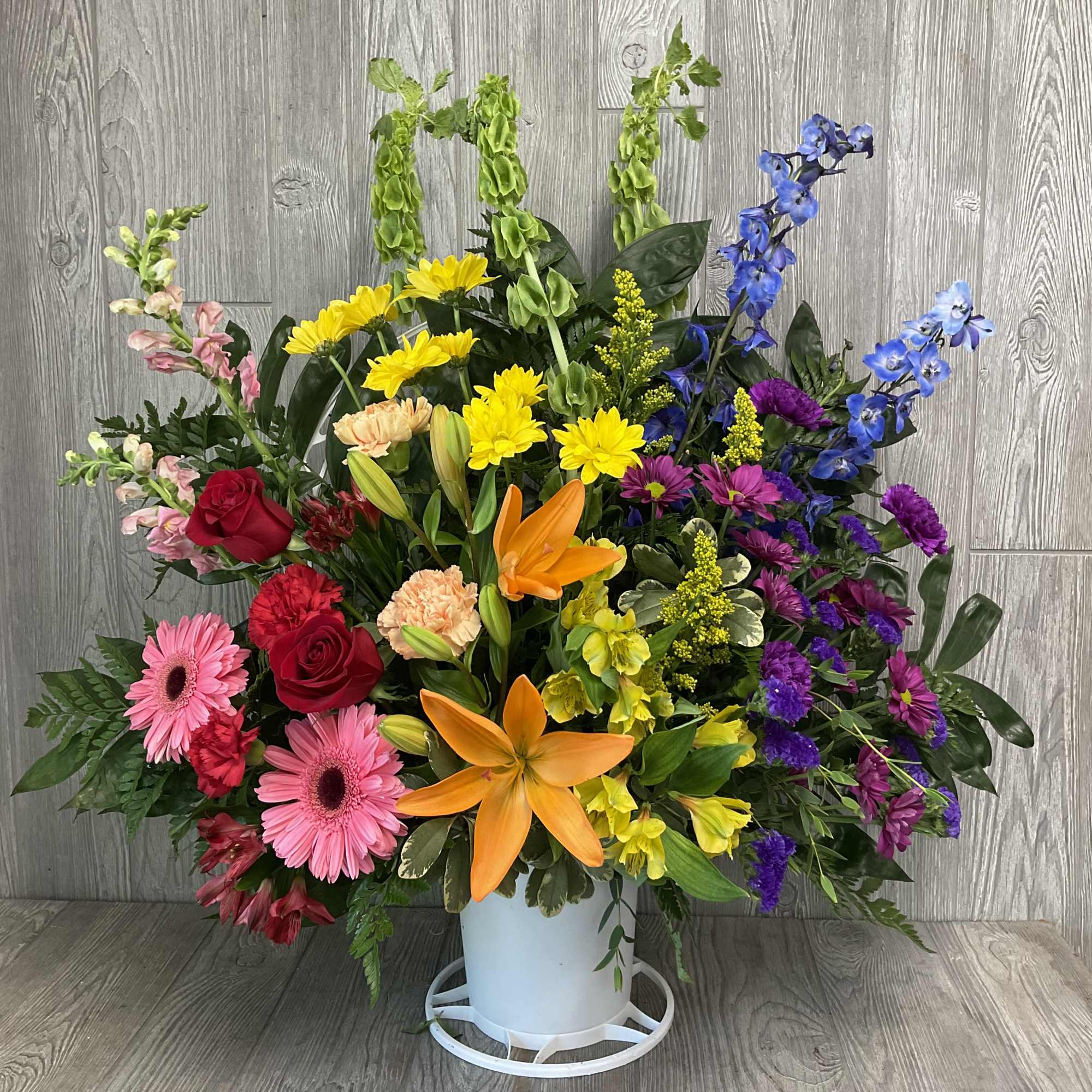 Tall mixed arrangement of colorful lilies, roses, daisies, and other blooms in a white container.