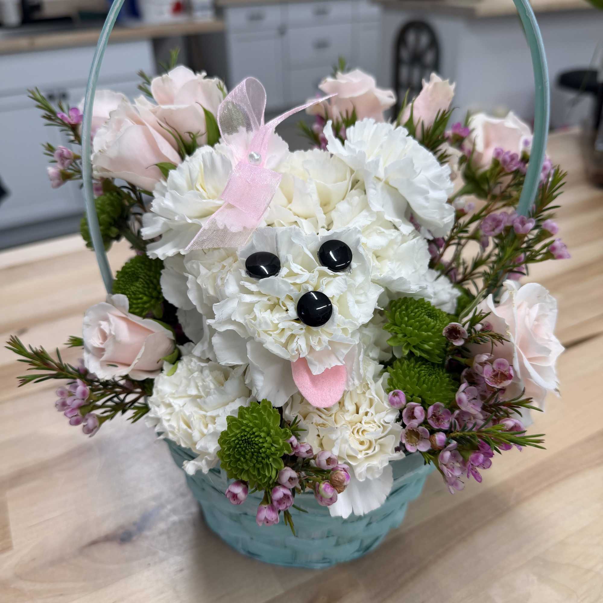 Basket arrangement with a white carnation dog surrounded by pink roses and small purple flowers.