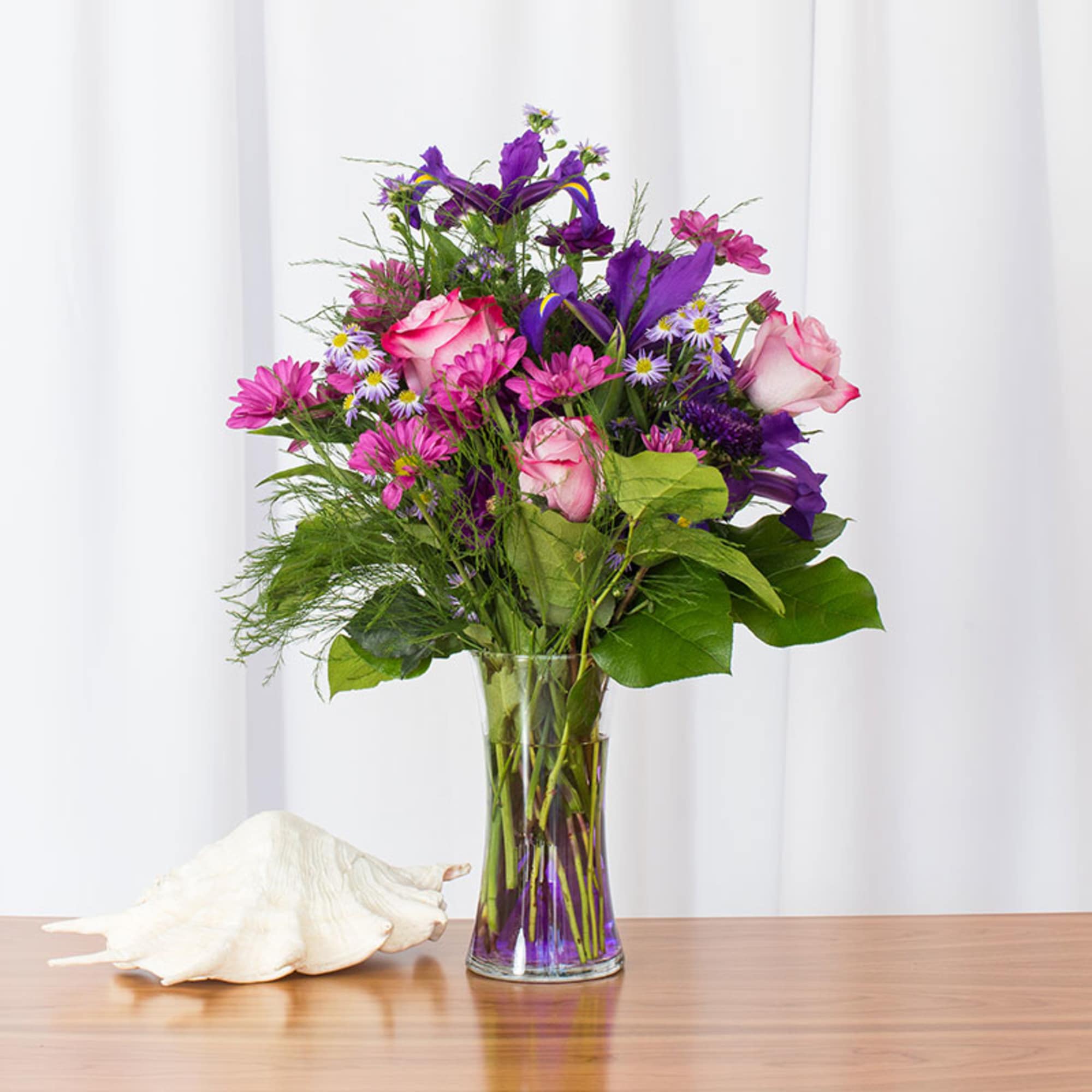 Tall glass vase of pink roses, purple irises, and mixed purple daisies beside a white seashell.