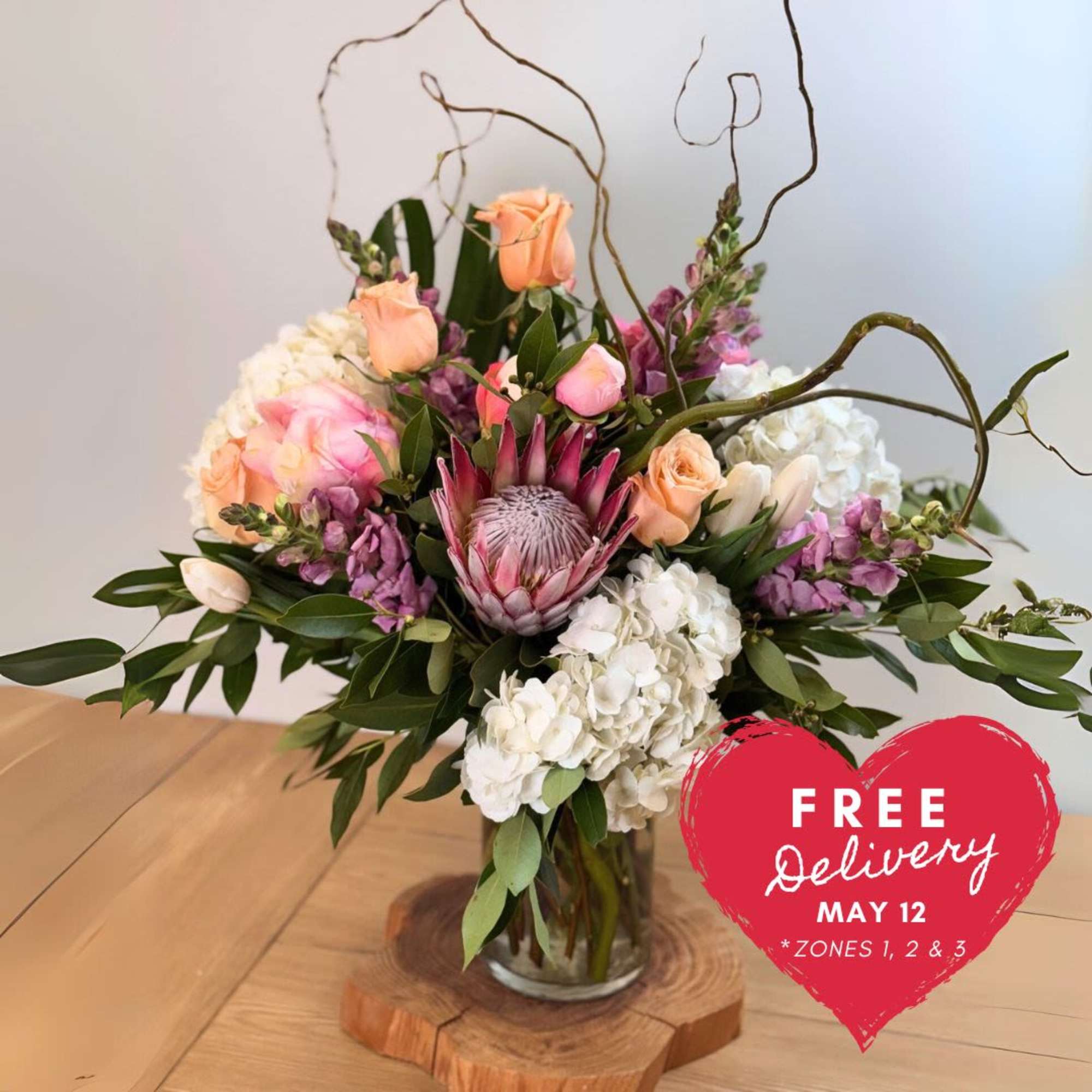 Mixed bouquet of peach roses, white hydrangeas, and a pink protea in a glass vase on a wood base.
