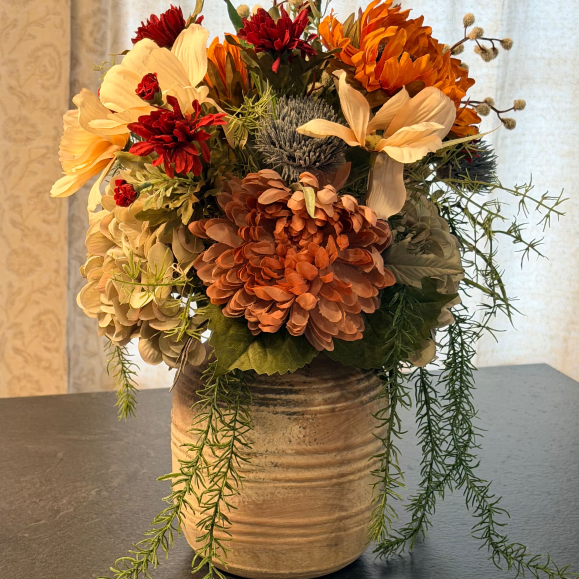 Autumn-toned mixed flower arrangement in a rustic ceramic vase with trailing greenery.