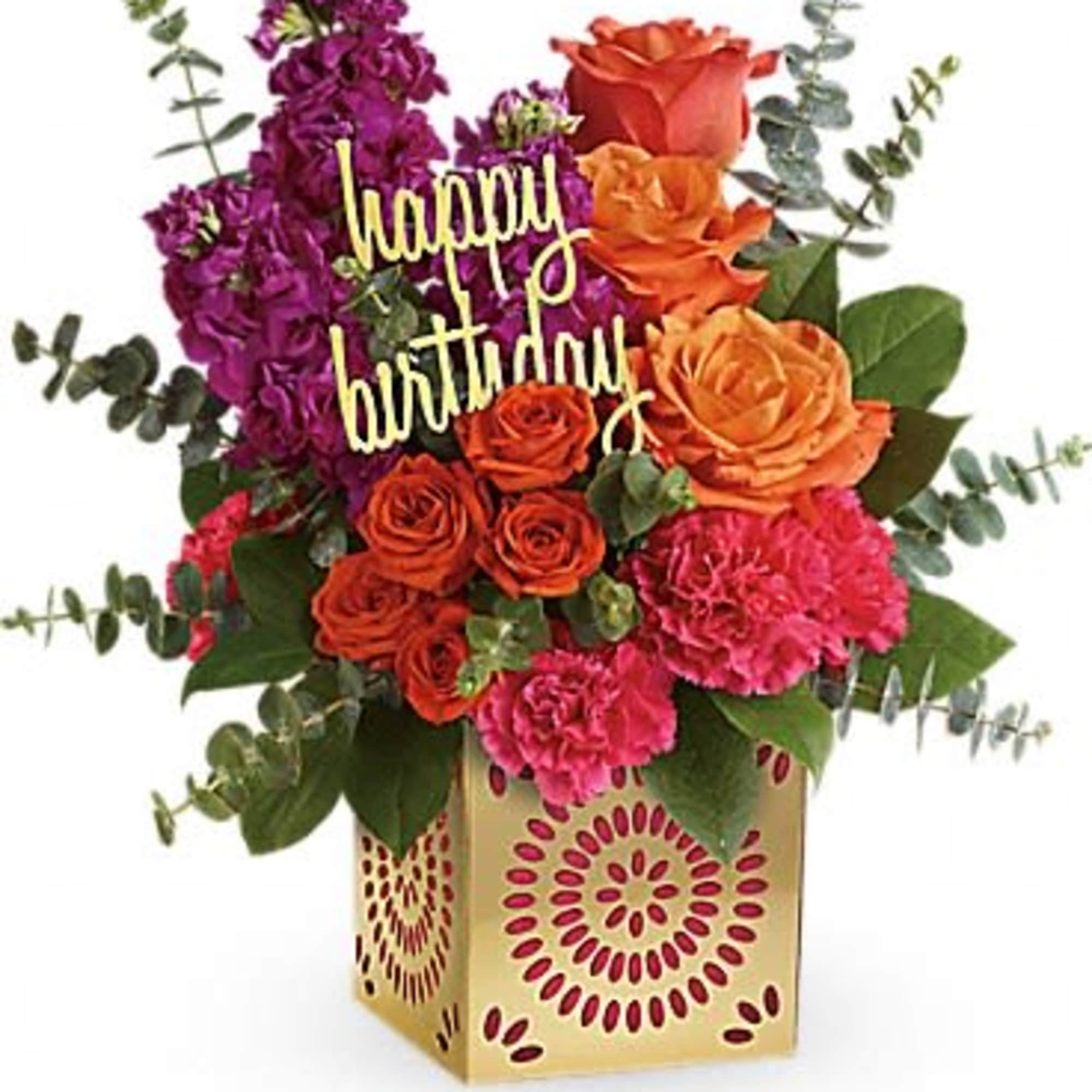 Bright arrangement of orange roses, pink carnations, and purple blooms in a gold patterned cube with a happy birthday sign