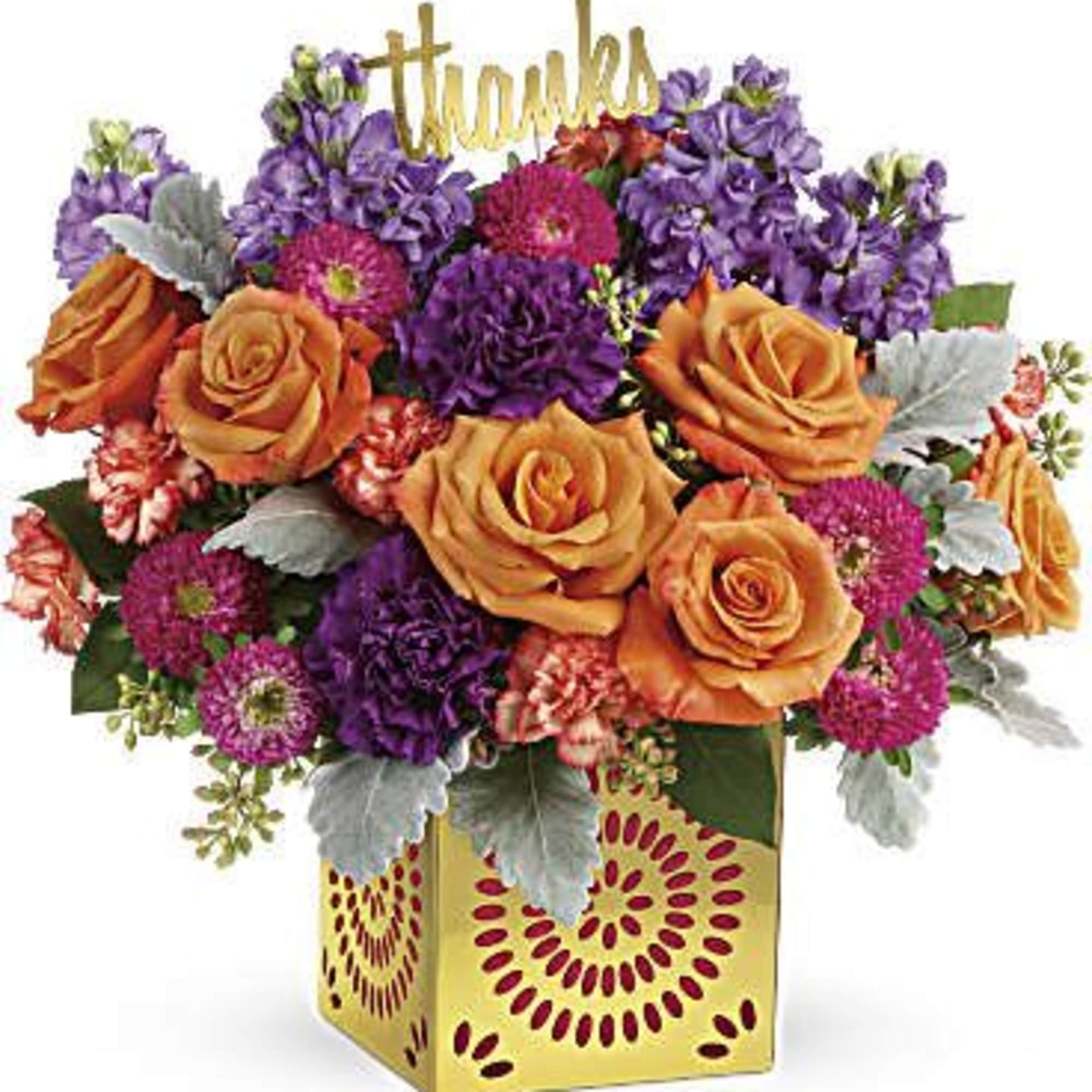 Colorful arrangement of orange roses and purple blooms in a gold cube vase with a thanks sign.