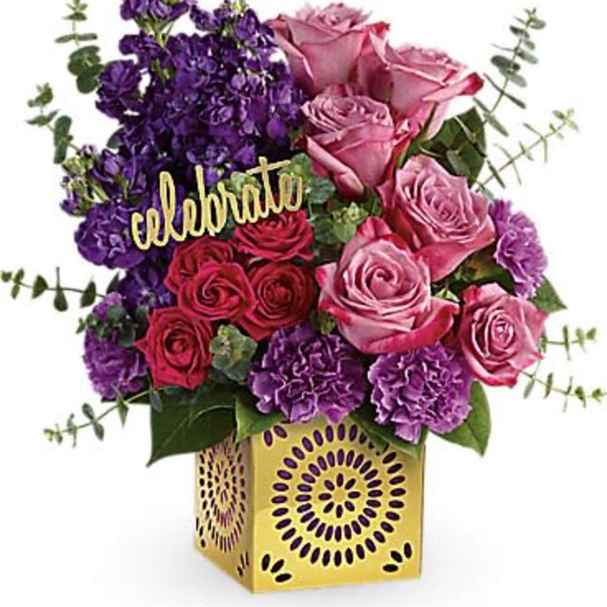 Compact arrangement of pink roses and purple flowers in a gold cube vase with a celebrate pick.