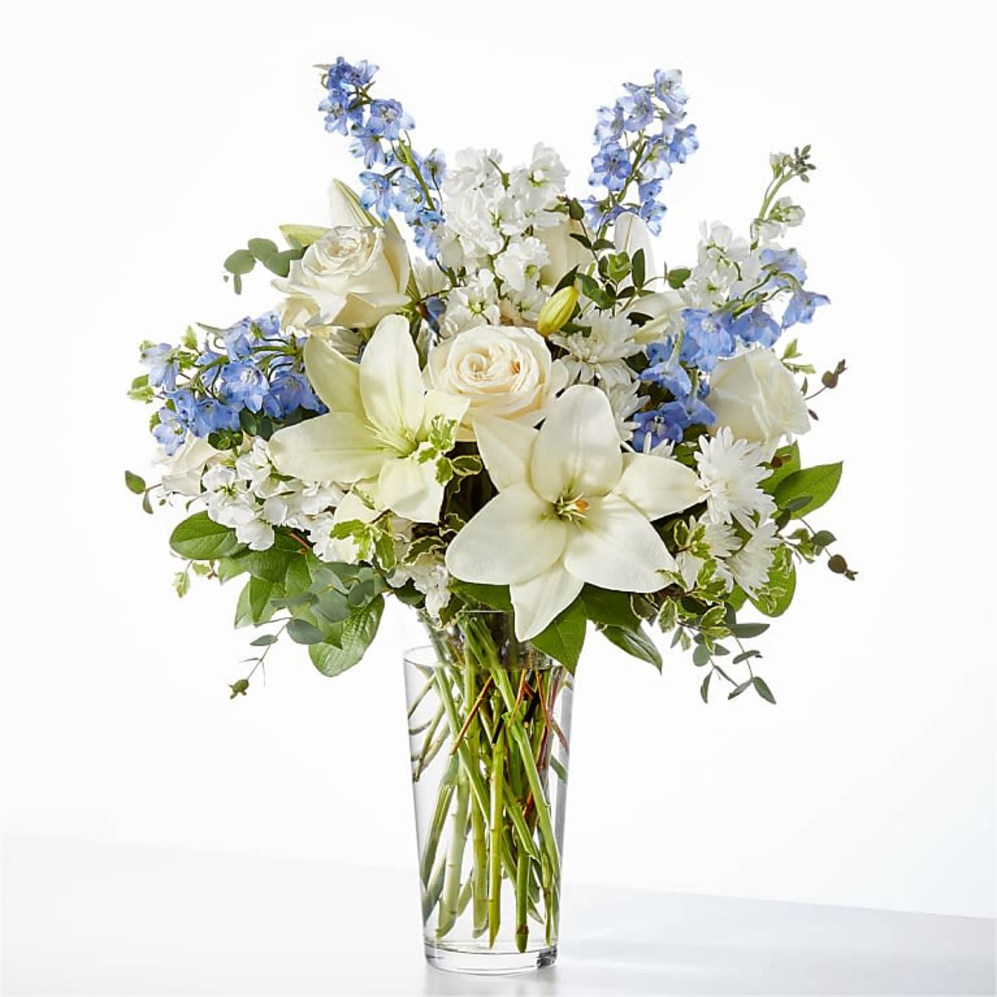 Tall glass vase of white lilies, roses, and mixed white flowers with soft blue accents.