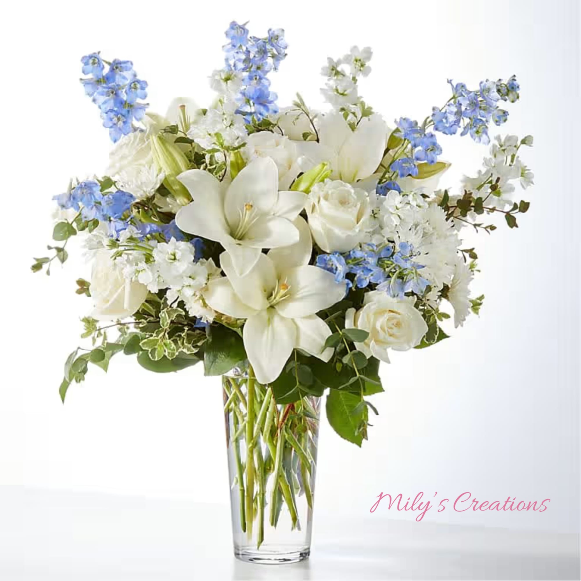 Tall clear vase of white lilies and roses with blue delphinium and white accent flowers.