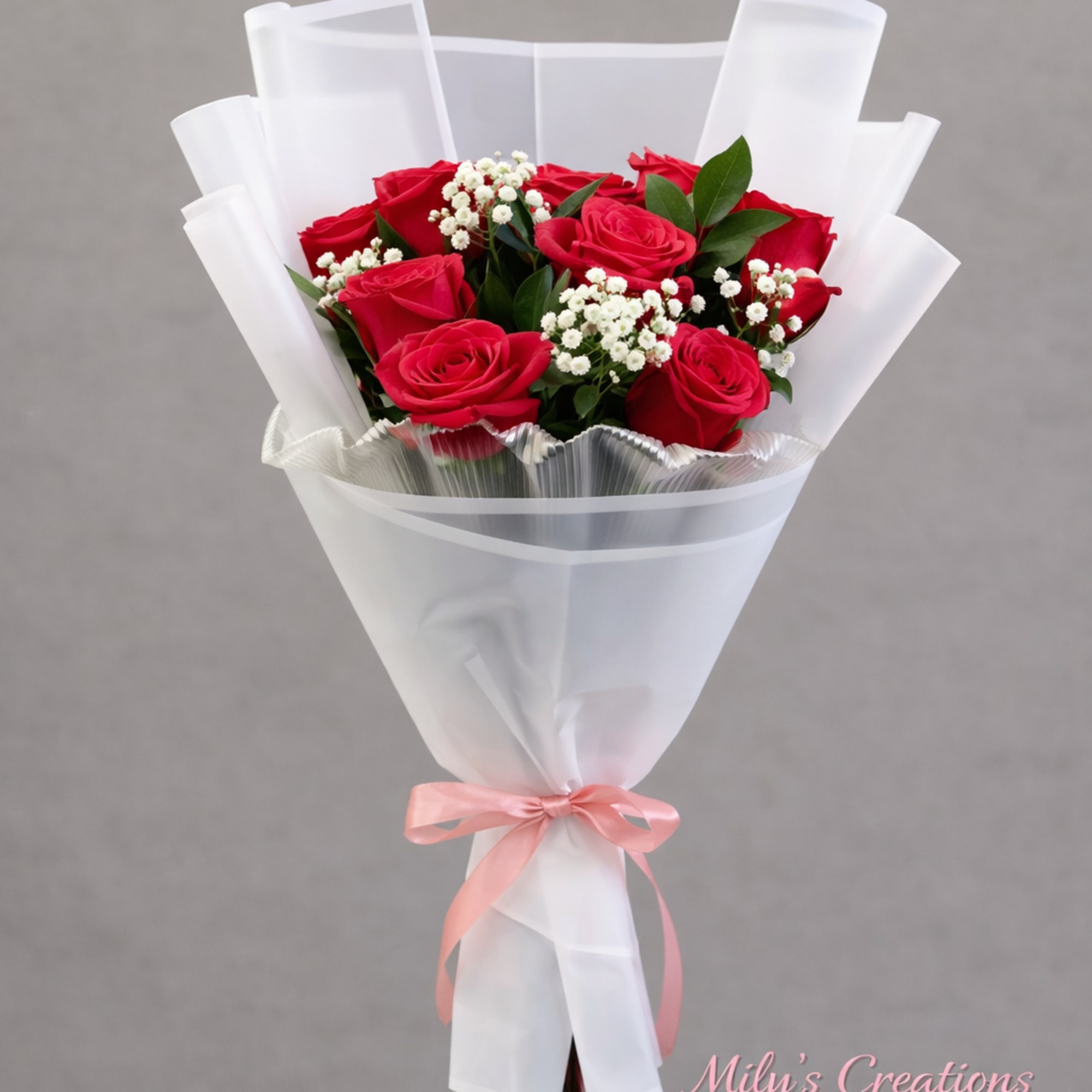 Bouquet of red roses with small white filler flowers wrapped in white paper and tied with a pink ribbon.