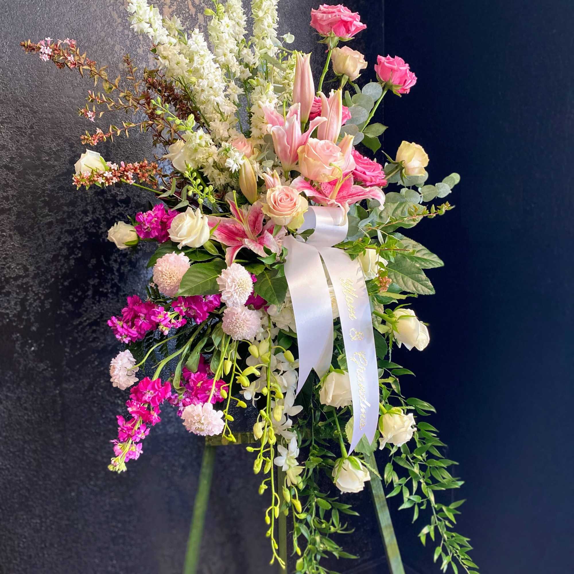 Tall pink and white standing floral arrangement with lilies, roses, and ribbon on a green easel.