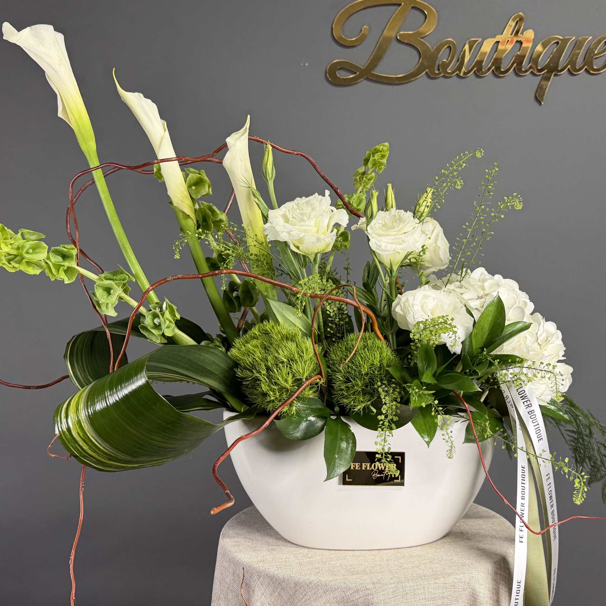 Modern white and green arrangement with calla lilies, roses, and sculpted foliage in a white ceramic bowl