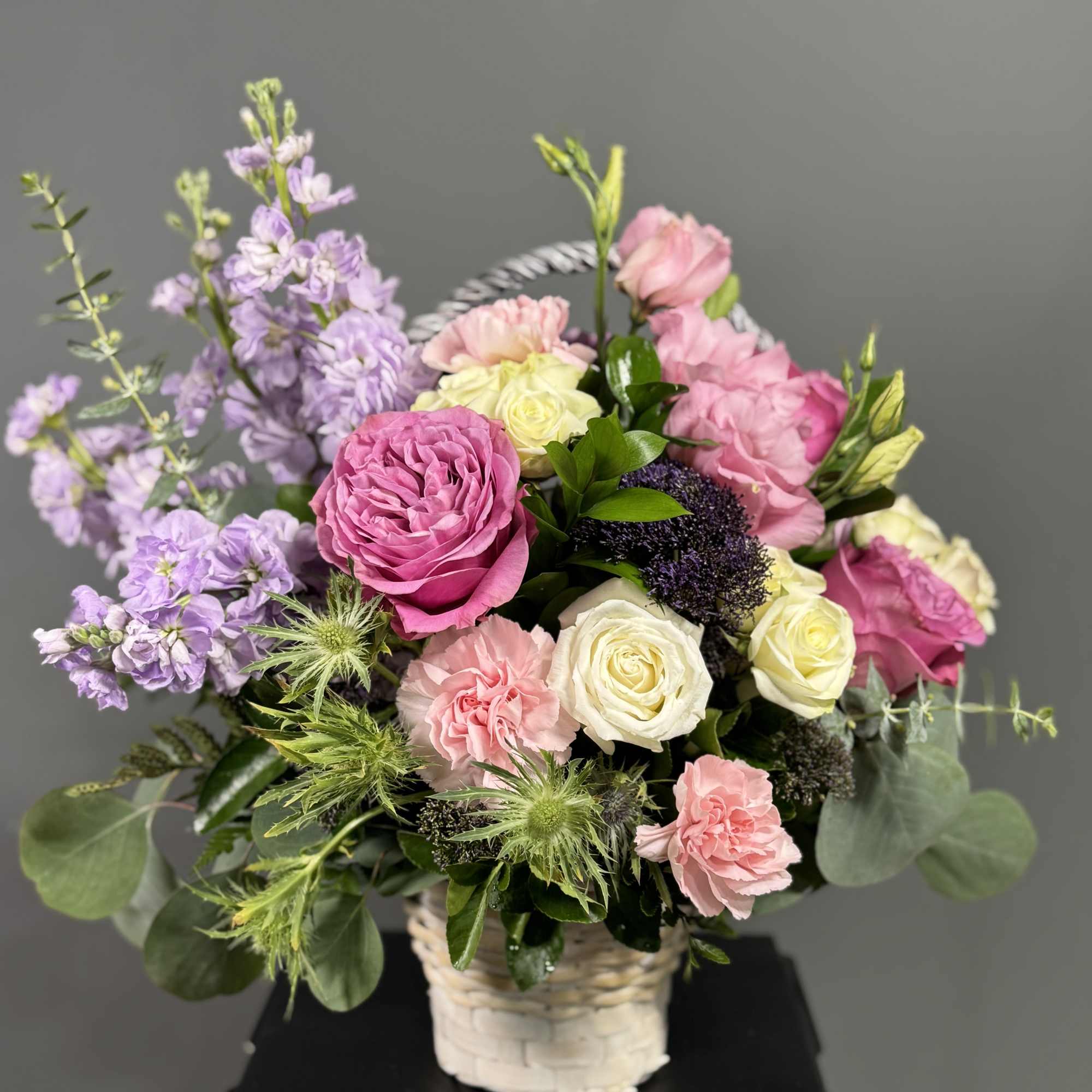 Basket arrangement of pink and cream roses with lavender stock and green thistle blooms