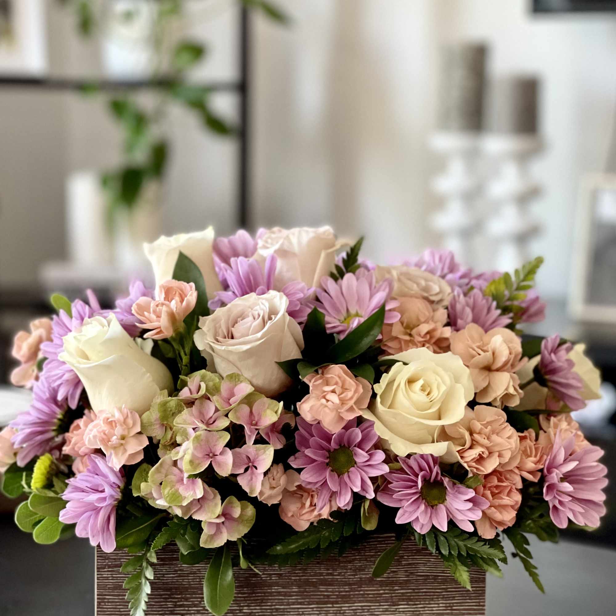 Low rectangular arrangement of cream roses, peach carnations, and lavender daisies in a wood box.