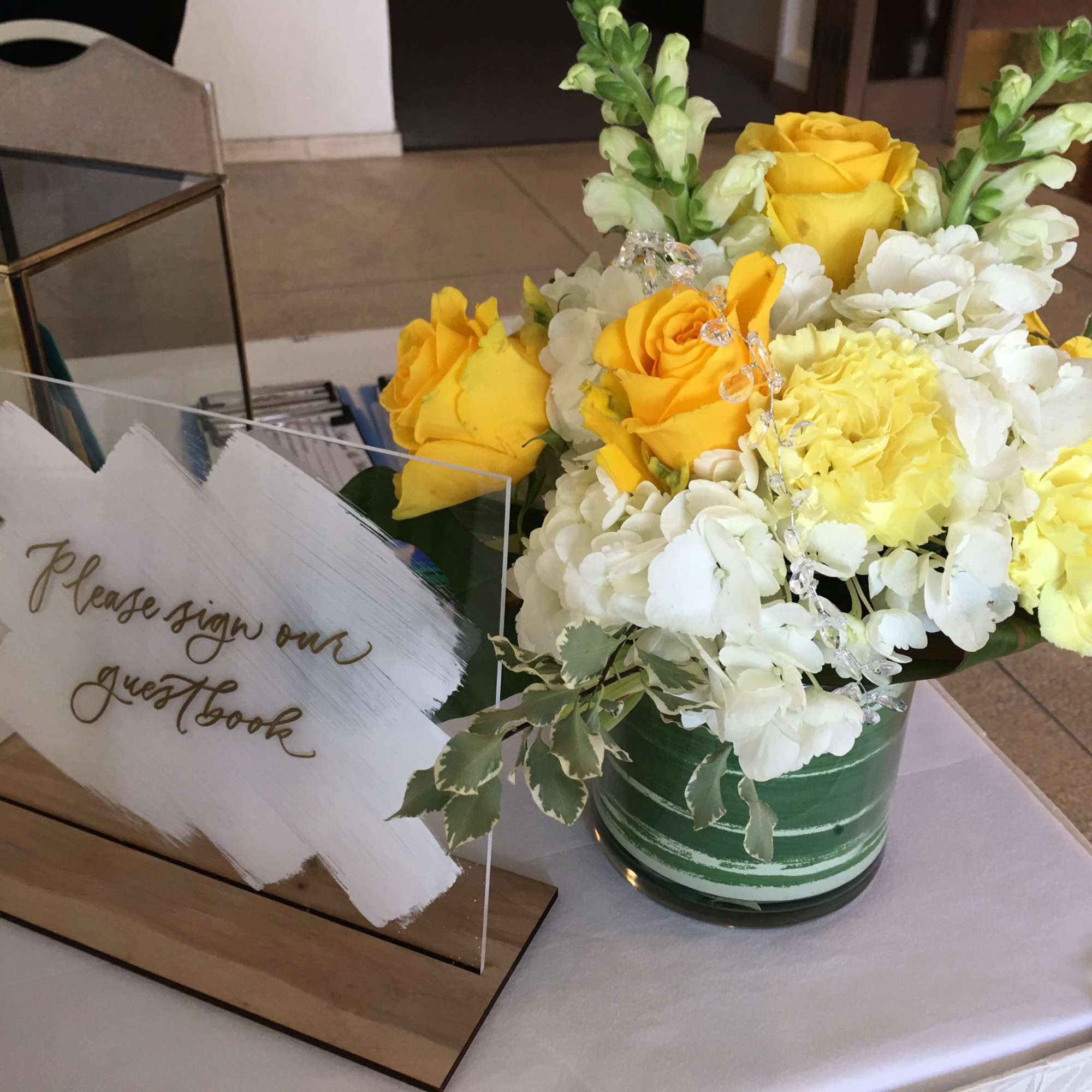 Yellow and white flower arrangement with roses and snapdragons in a green vase beside an acrylic guestbook sign