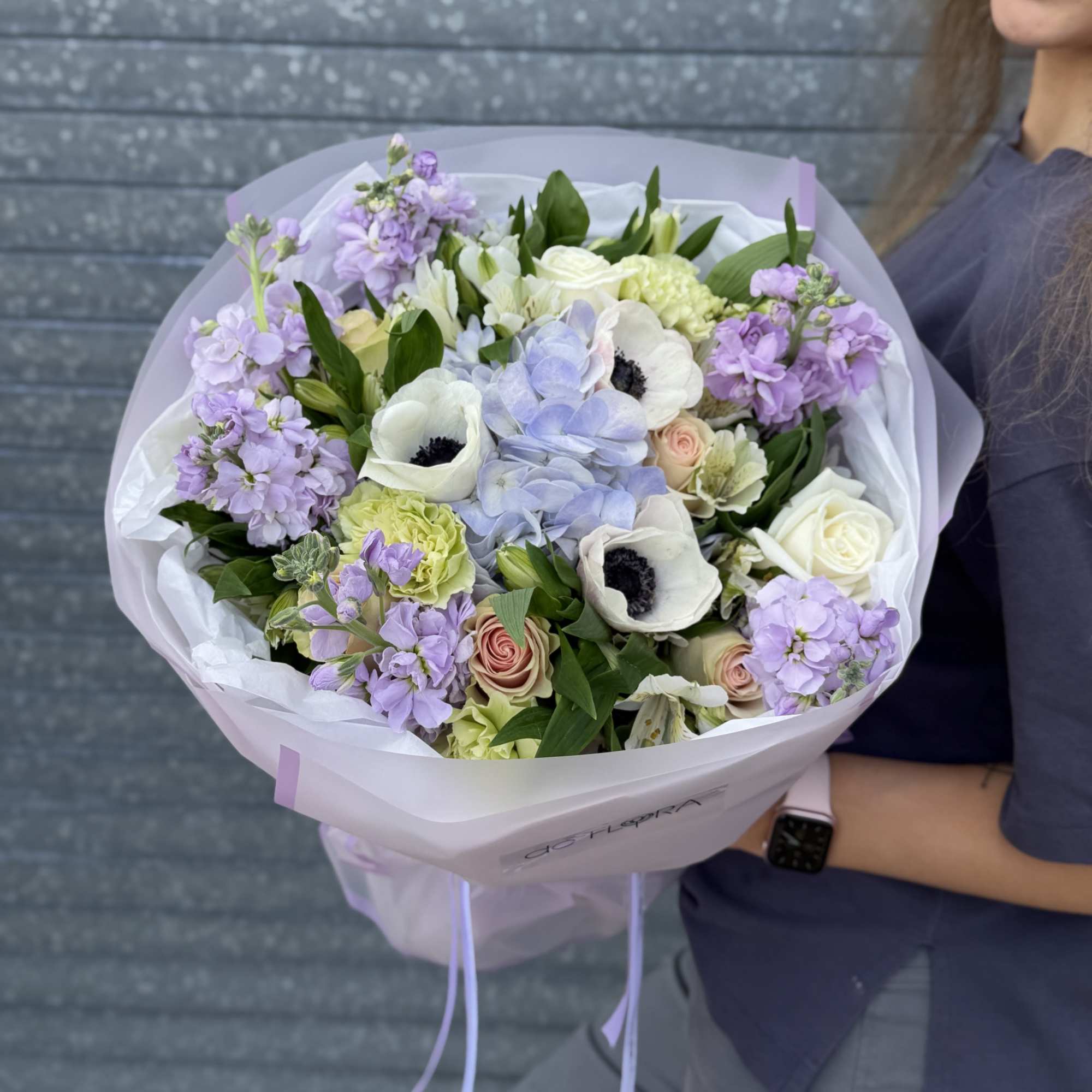 Large pastel bouquet with white anemones, blue hydrangeas, roses, and lavender stock in white wrap held by a person.