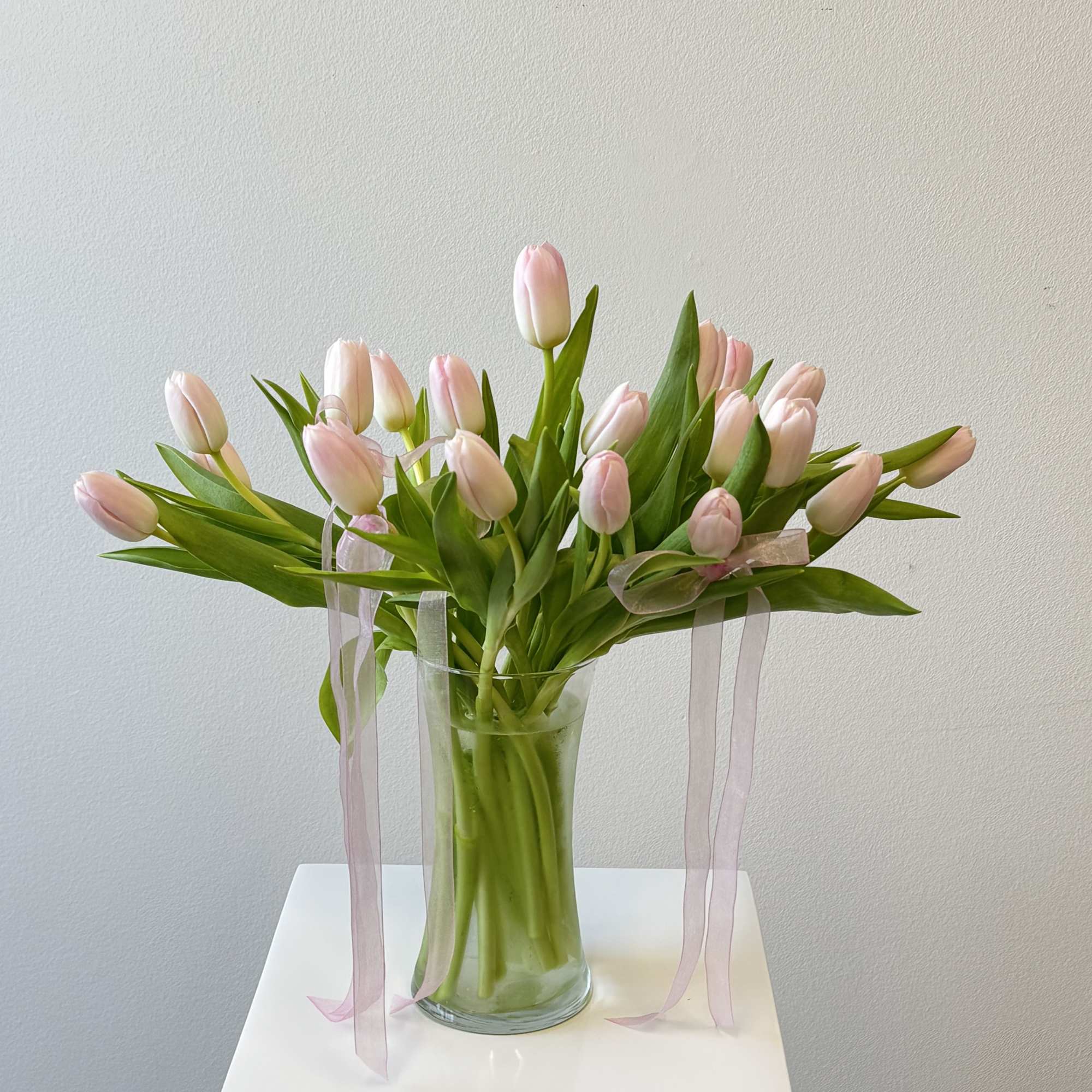 Arrangement of pale pink tulips in a clear glass vase with trailing pink ribbons.