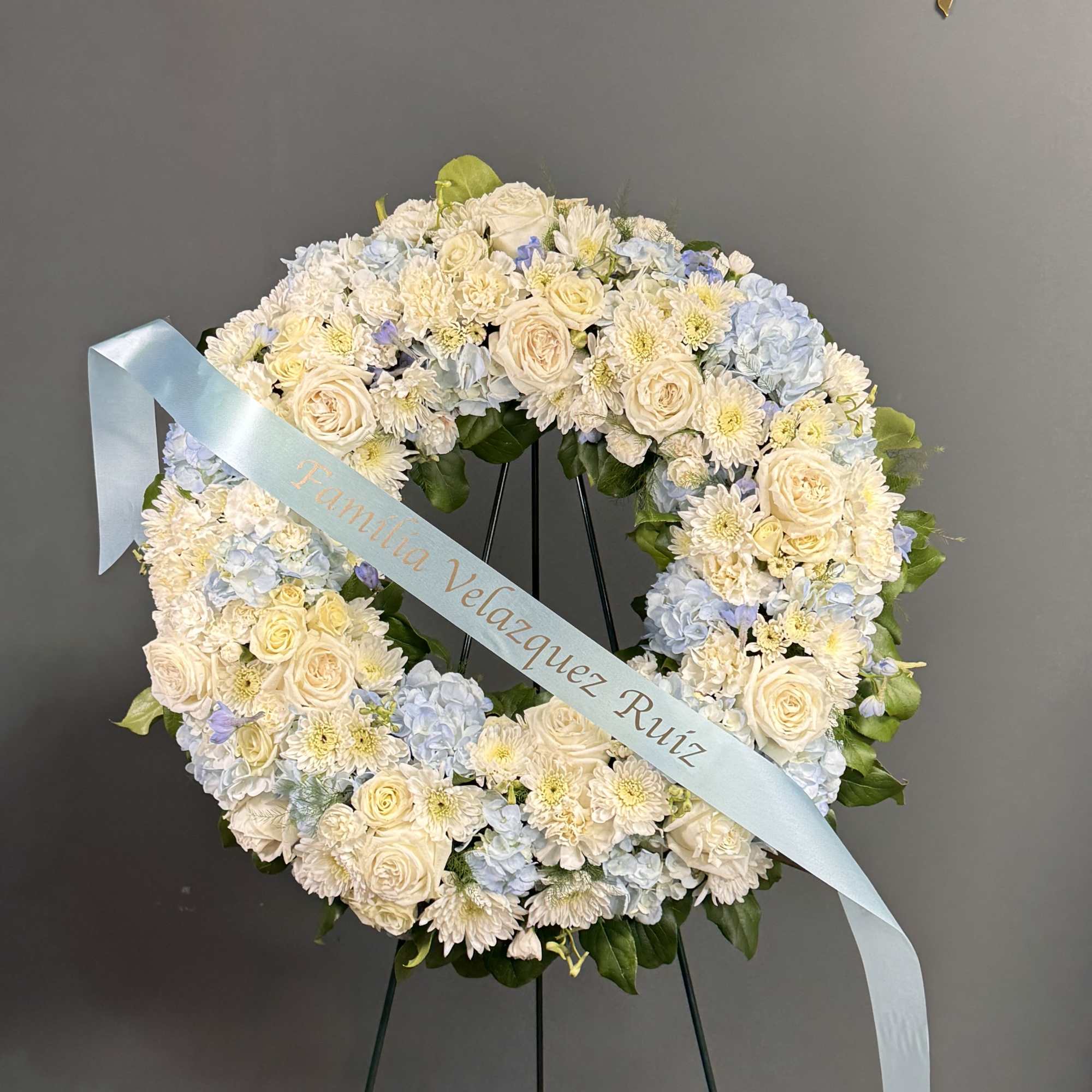 Standing round wreath of white roses, mums, and blue hydrangeas on an easel with a light blue ribbon.