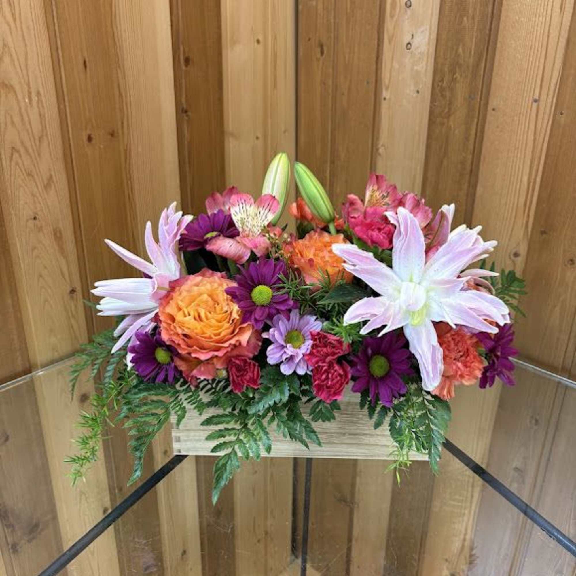 Low wooden box arrangement of pink lilies, orange rose, and bright mixed blooms with trailing foliage