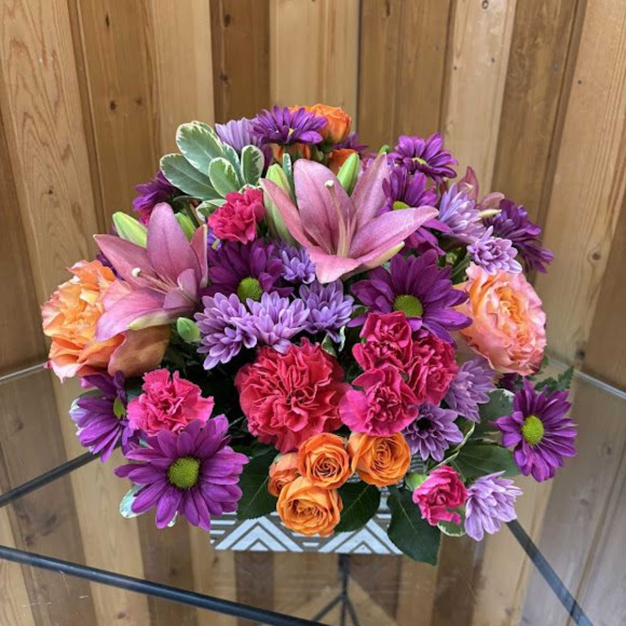 Bright mixed arrangement of purple mums, pink lilies, and orange roses in a patterned white vase.