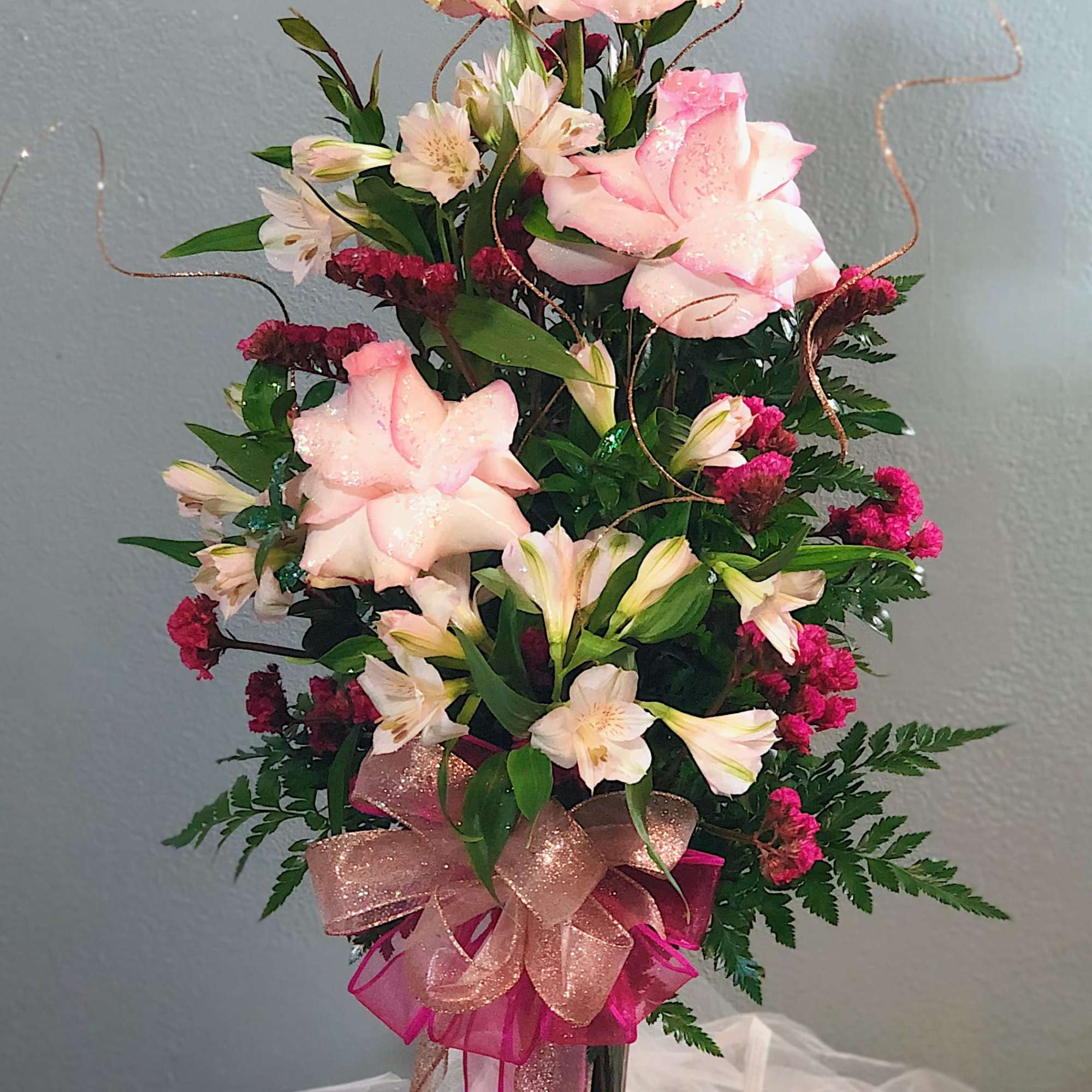 Tall pink and white rose and alstroemeria arrangement in a dark glass vase with glittery ribbons