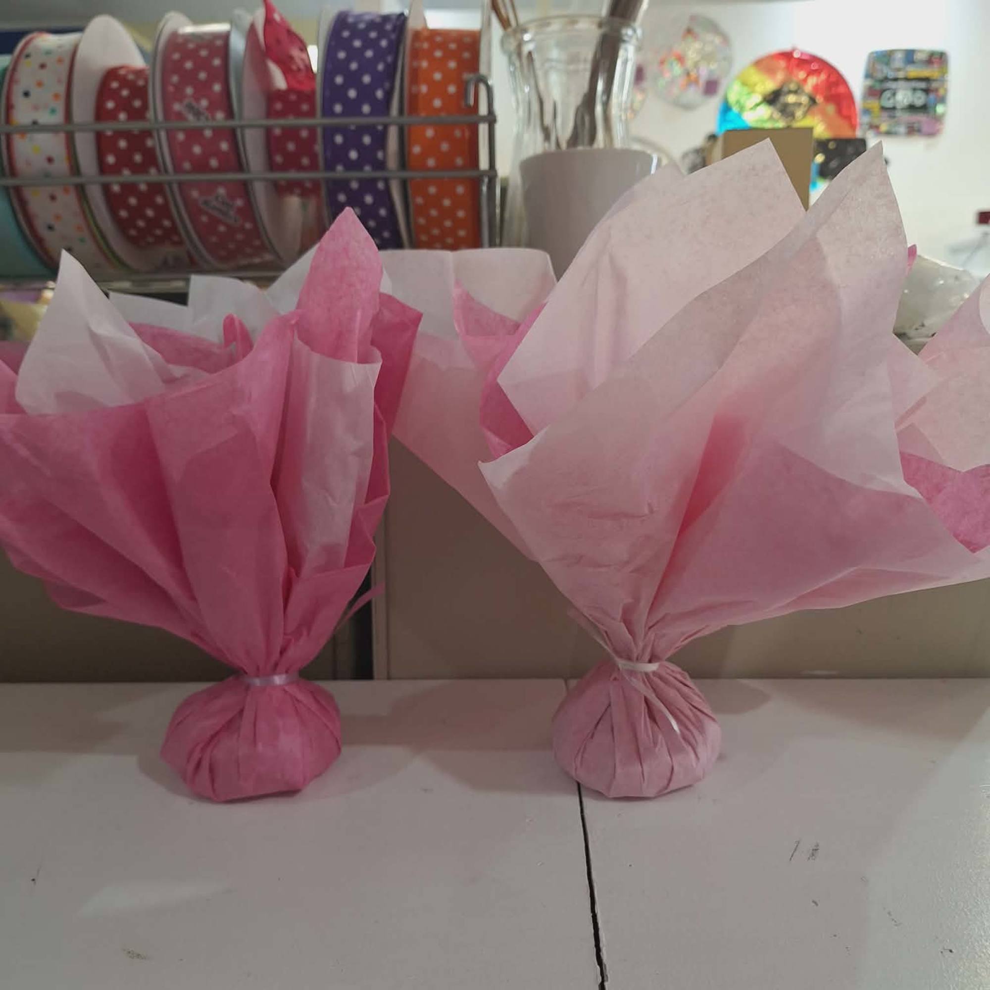 Two small items wrapped in pink tissue paper on a white shelf with colorful ribbon spools behind.
