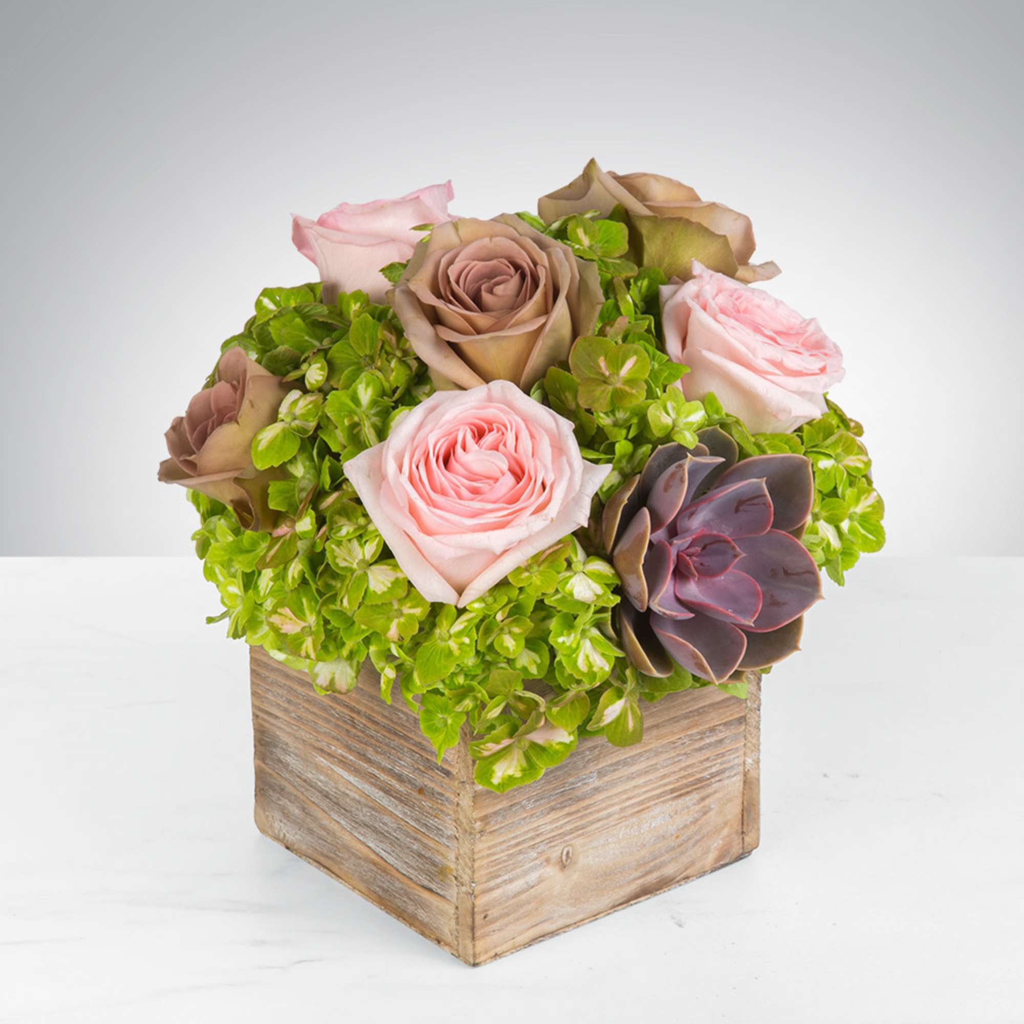 Compact arrangement of pink roses, green hydrangeas, and a succulent in a rustic wooden cube.