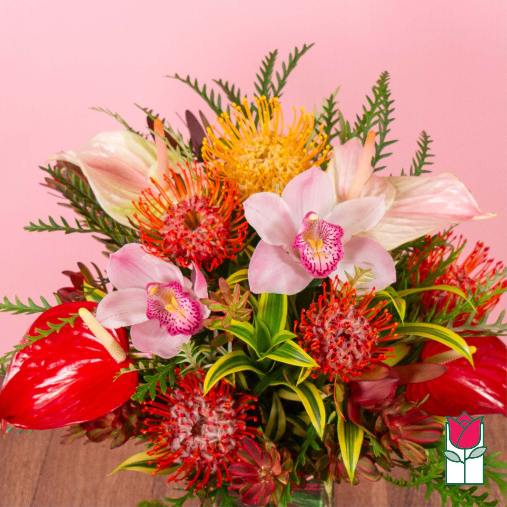 Tropical bouquet with pink orchids, red blooms, yellow pincushion protea and variegated foliage in a glass vase