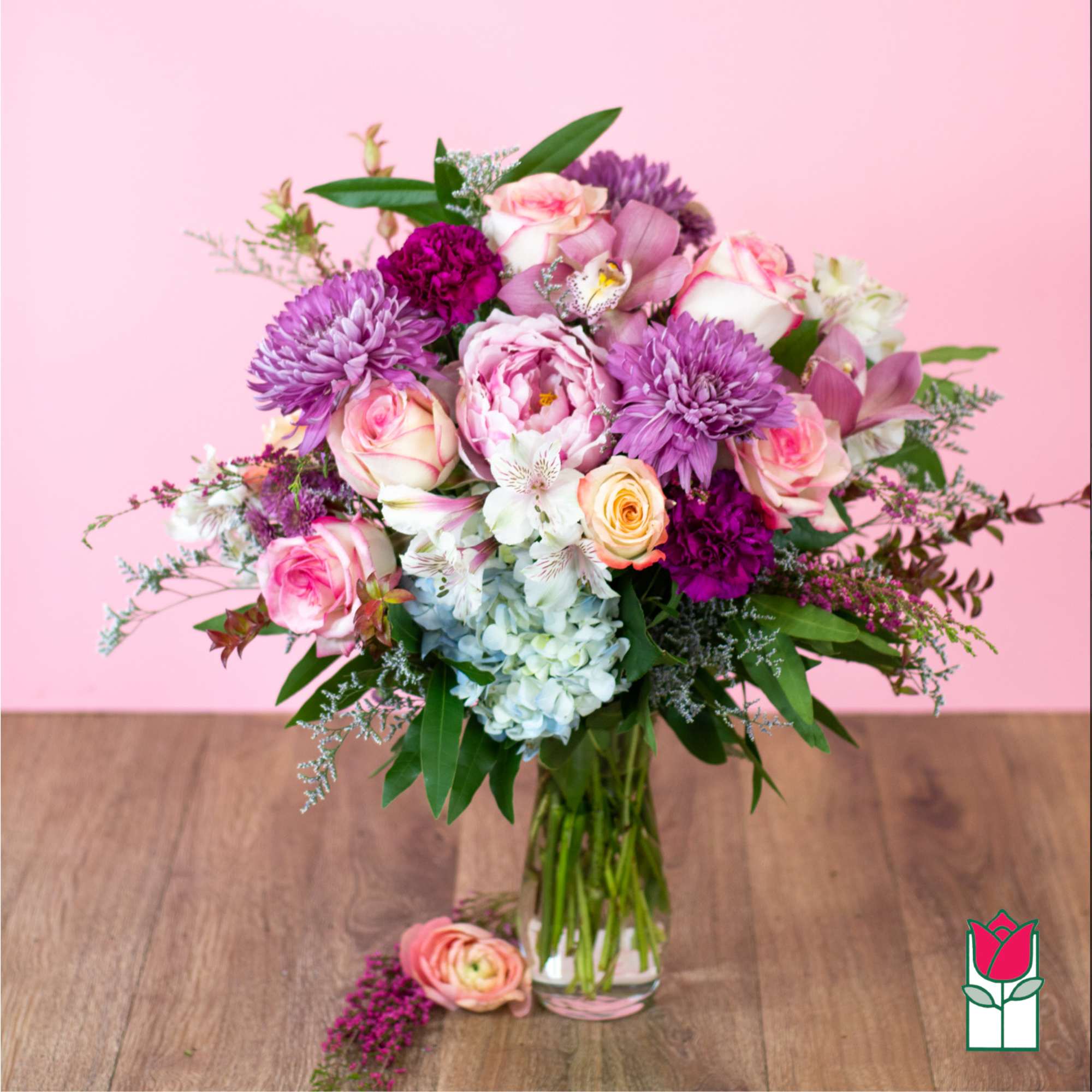 Vase arrangement of pink roses, purple mums, peonies, and blue hydrangea against a pink background