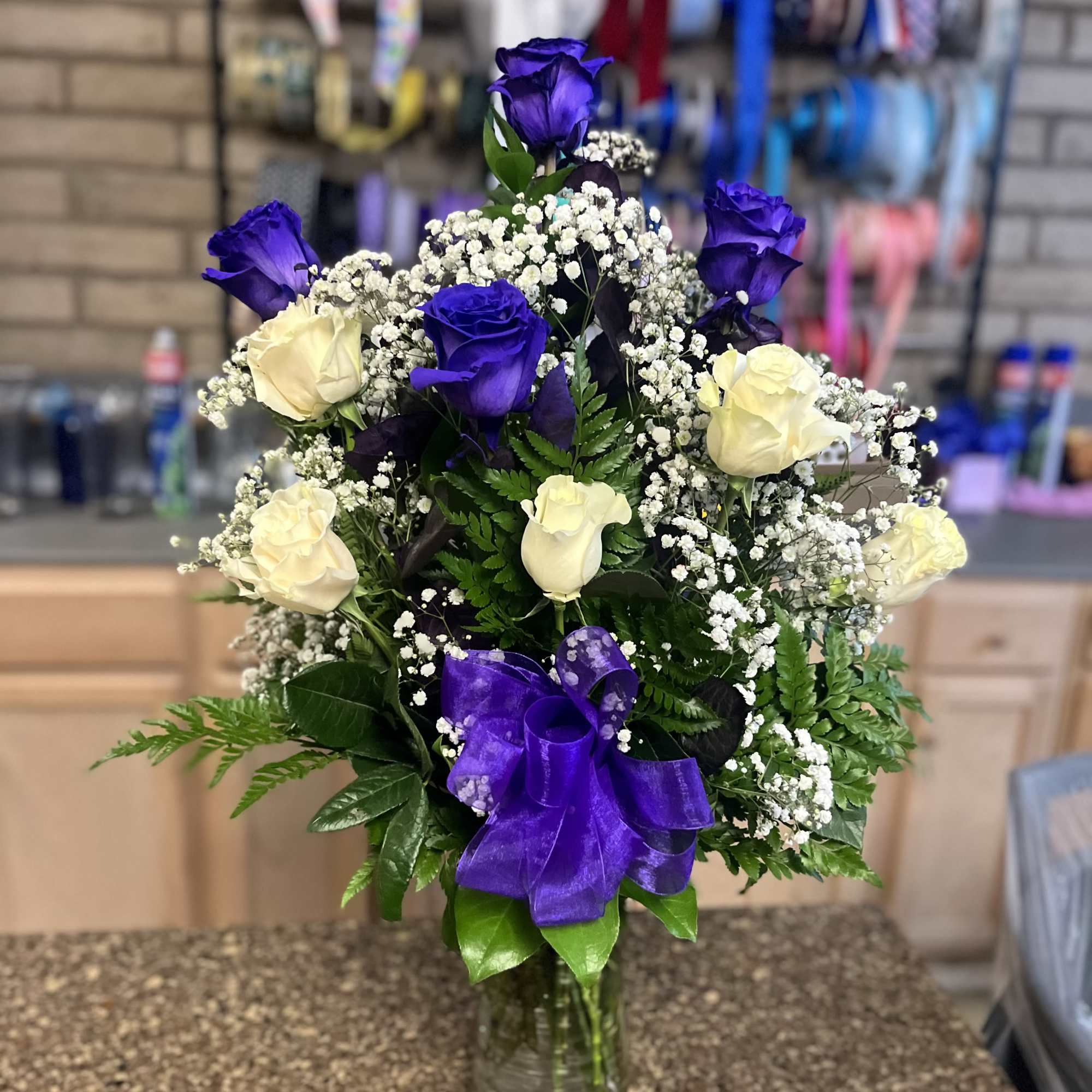 Tall arrangement of purple and white roses with baby's breath in a clear glass vase with a purple bow.